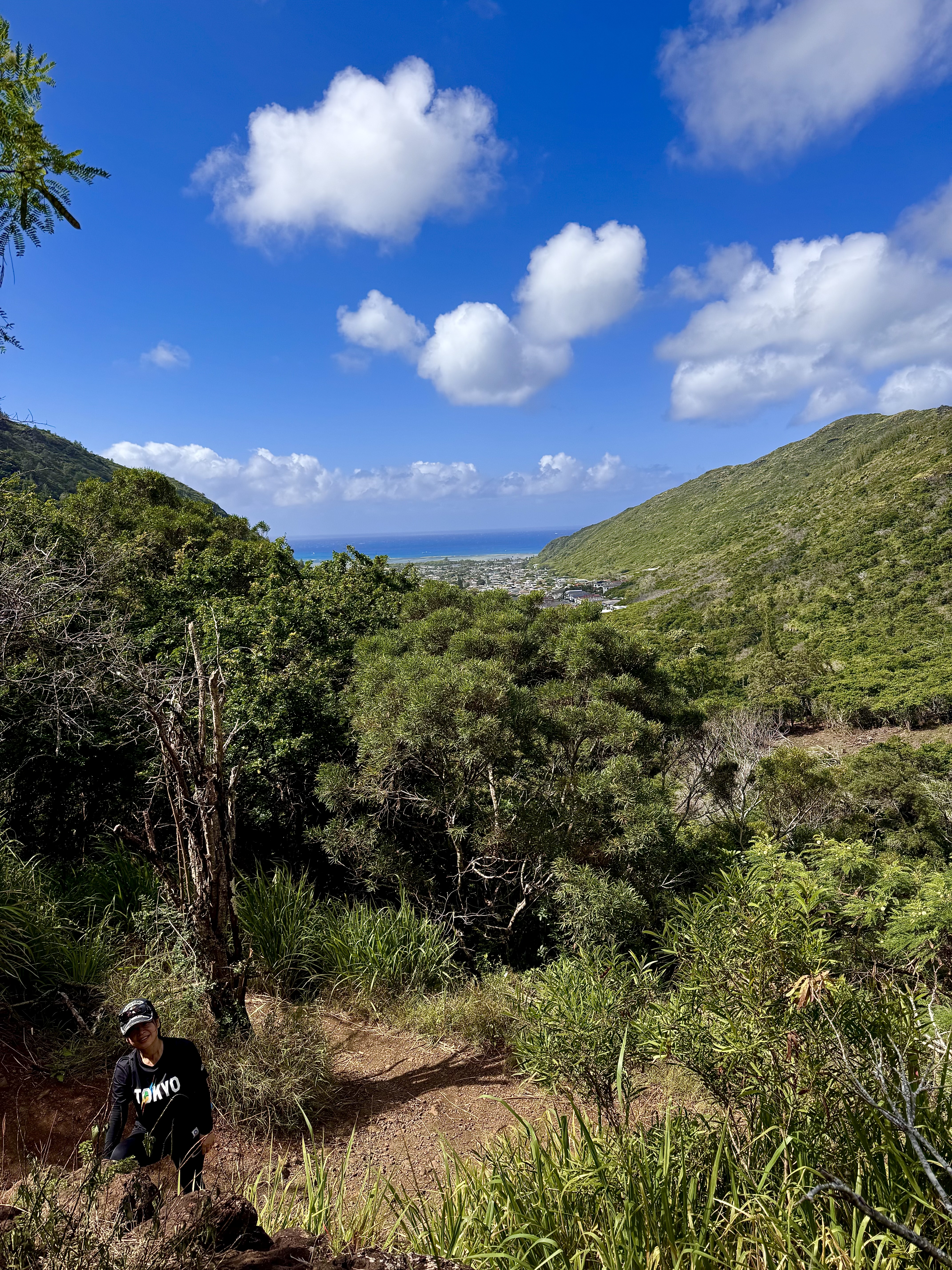 Ayumi in Tokyo shirt climbing up the trail with a sweeping valley and ocean view behind her