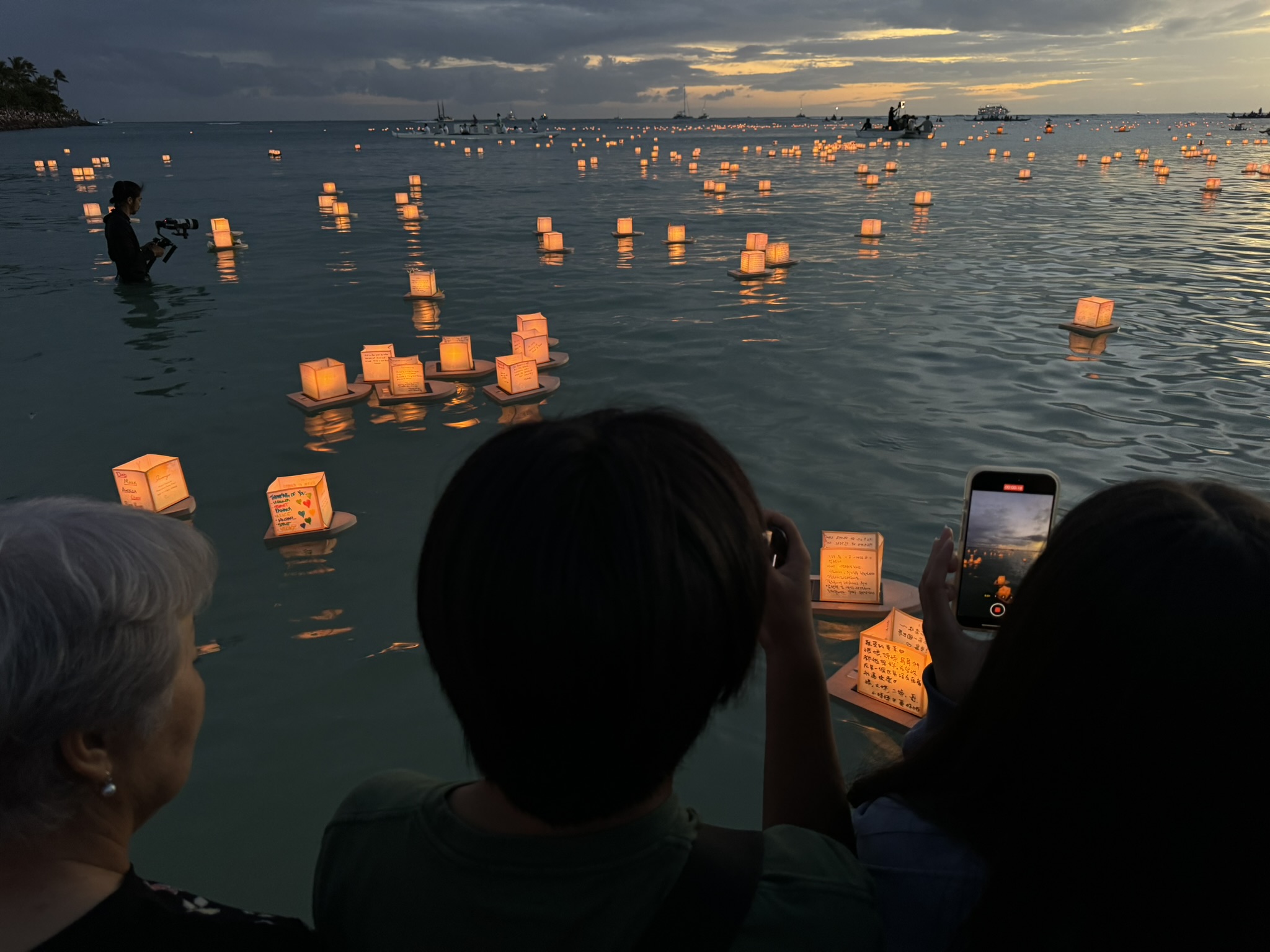 John's mother, Jace, and Maile at the water's edge watching their lanterns float out to sea, handwritten messages visible on the lanterns in the foreground