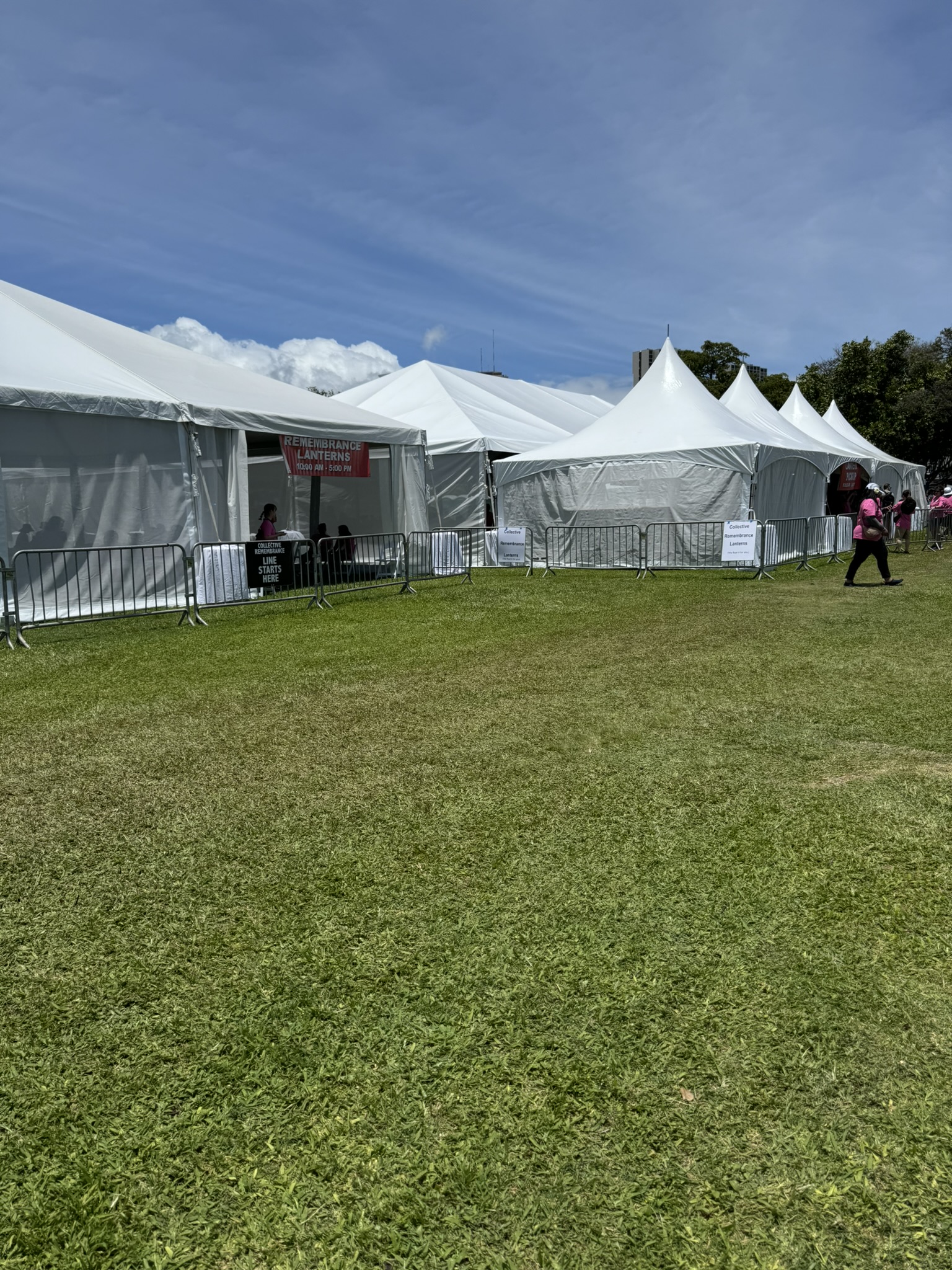 White event tents set up on the lawn at Ala Moana Beach Park, with a red 'Remembrance Lanterns' sign and a 'Collective Remembrance Line Starts Here' board