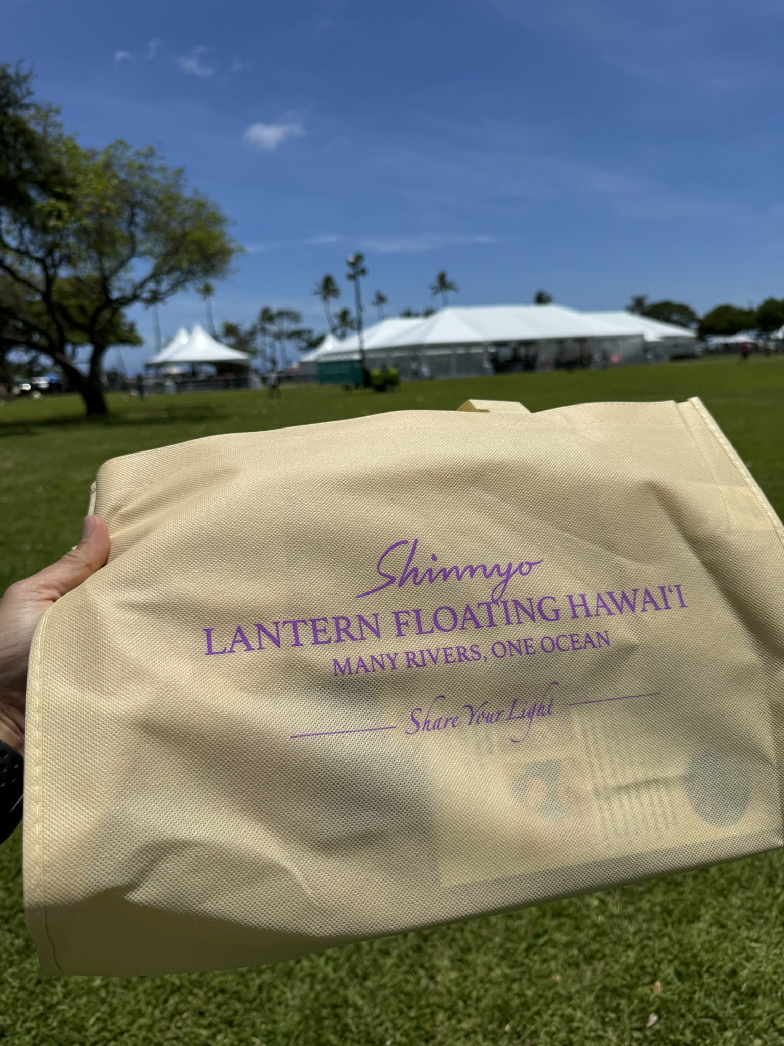 Hand holding the Shinnyo Lantern Floating Hawaii tote bag with the text 'Many Rivers, One Ocean' and 'Share Your Light'