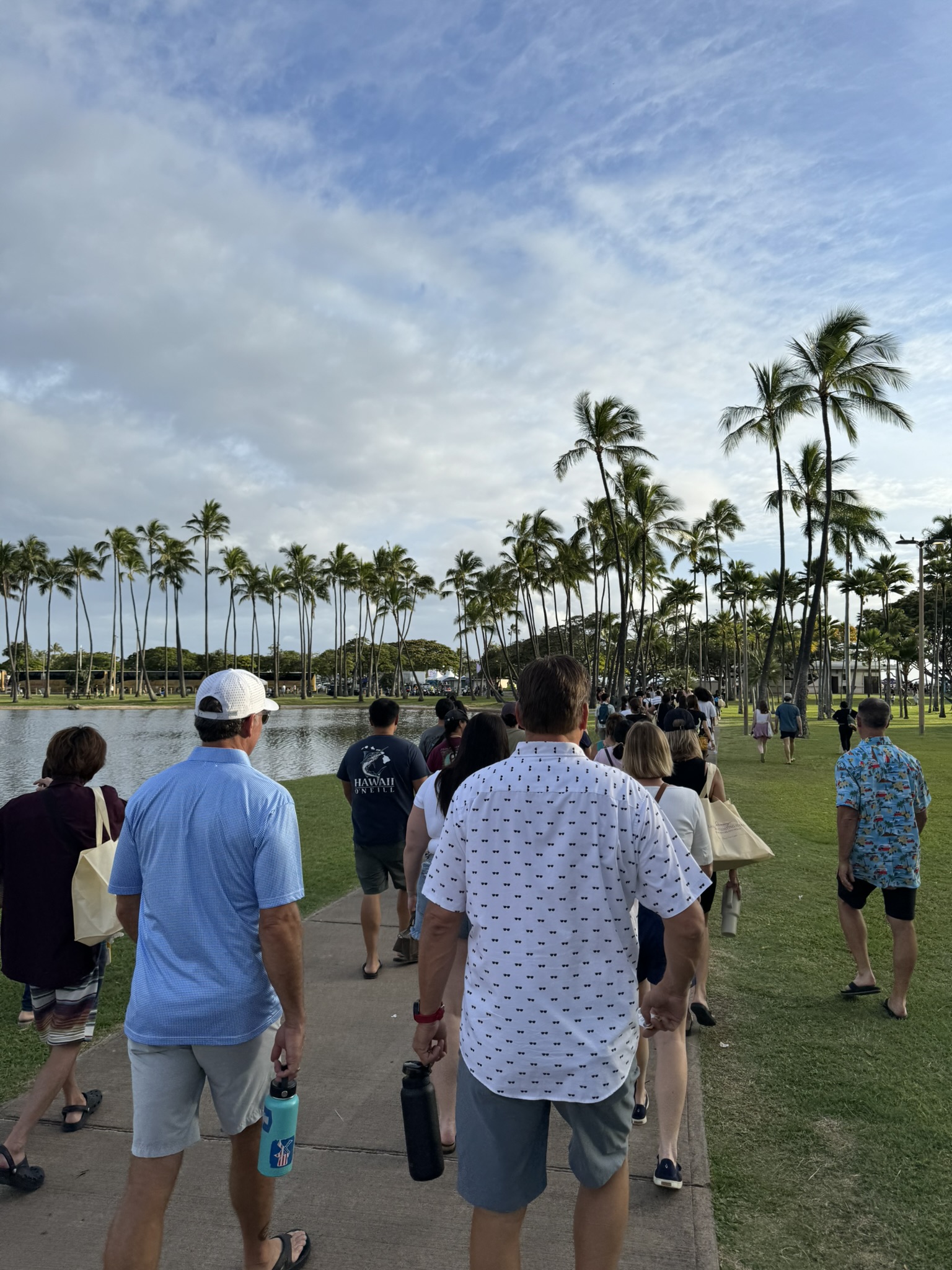 Jace walking in the foreground along the lagoon path toward Ala Moana Beach, with tall coconut palms lining the route and a crowd of people moving in the same direction