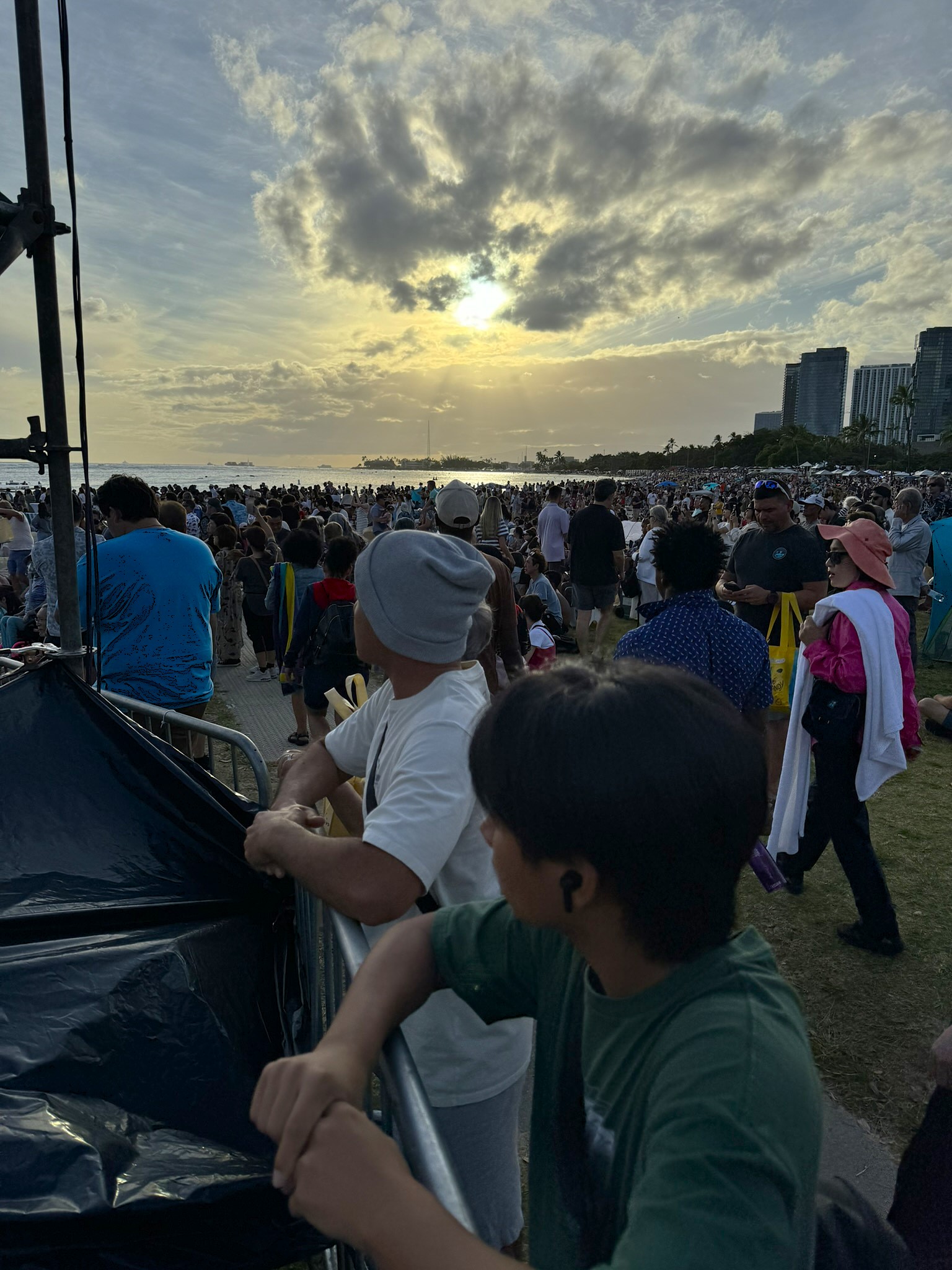 A massive crowd filling the beach and lawn at Ala Moana as the sun sets behind Honolulu's high-rise skyline