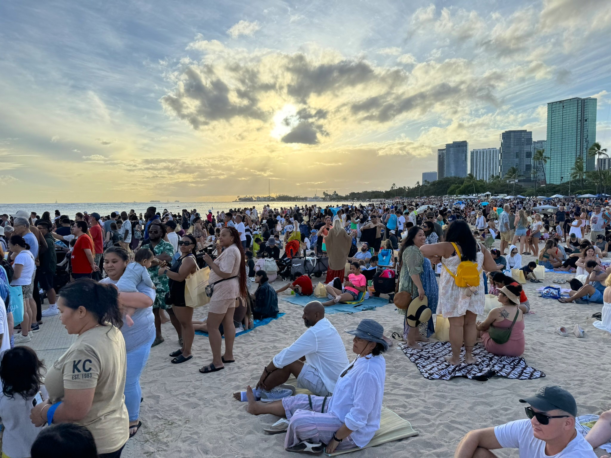 Thousands of people on the sand at Ala Moana Beach at golden hour, some sitting on blankets, others standing, with the Honolulu skyline visible along the waterfront