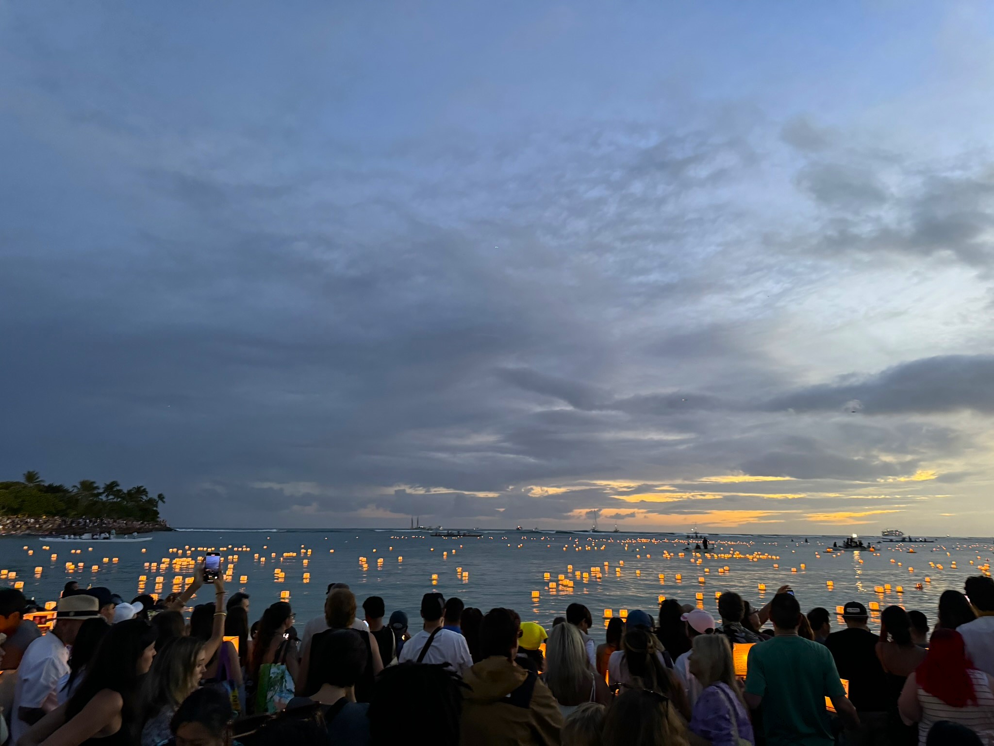 Thousands of glowing paper lanterns drifting out across the dark water at Ala Moana Beach at dusk, Honolulu skyline visible in the distance