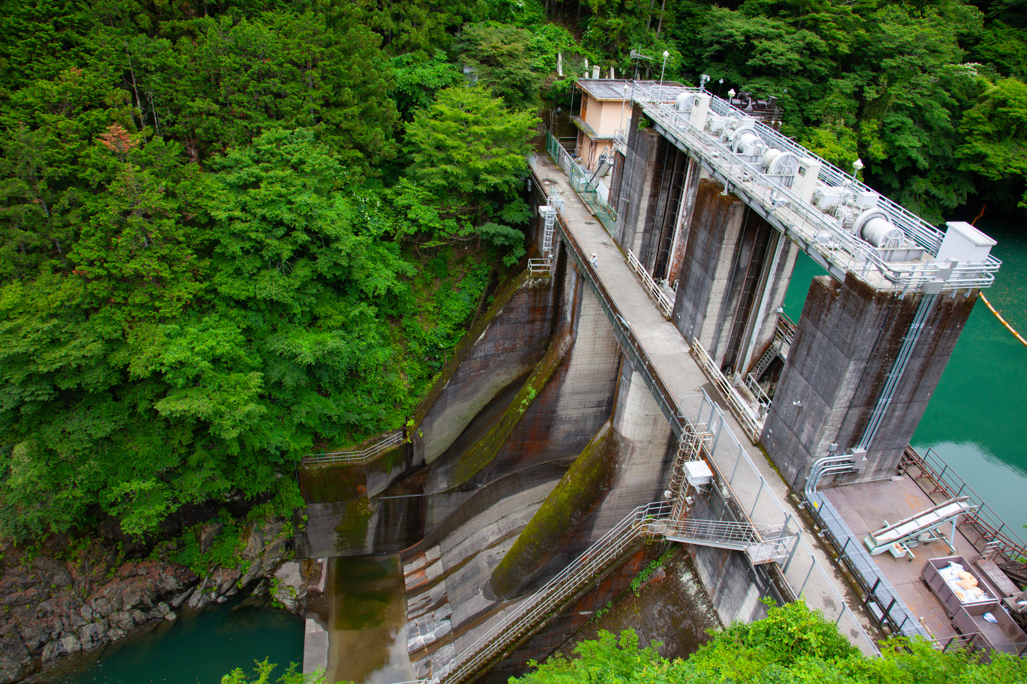 The Shiromaru Dam on the Tama River seen from above — a concrete dam and turbine building set into steep forested slopes, green reservoir water on the right and a mossy spillway channel below