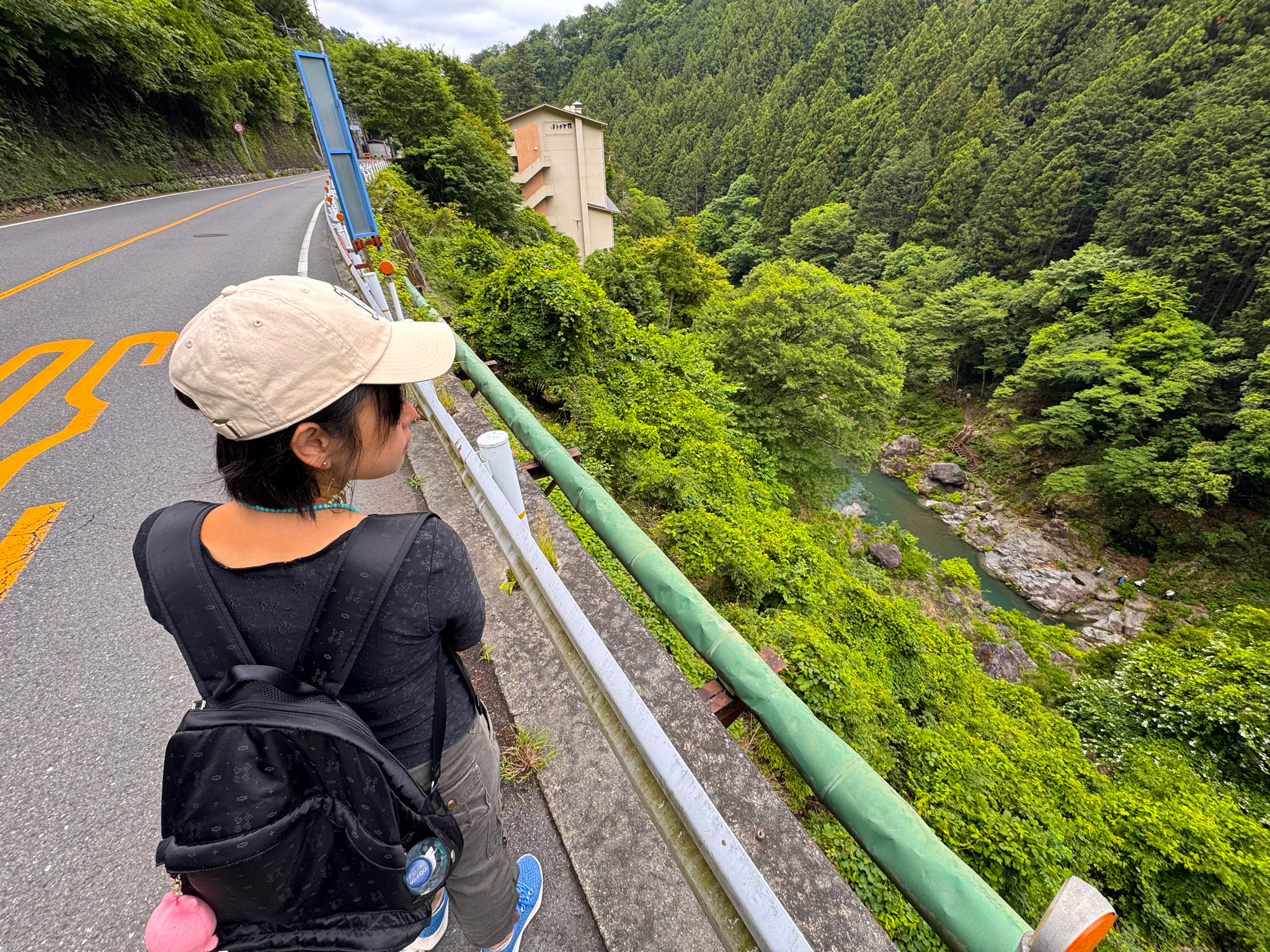 Ayumi walking back along the narrow mountain road above the gorge, a green water pipe and guardrail beside her, the river valley dropping away on the right