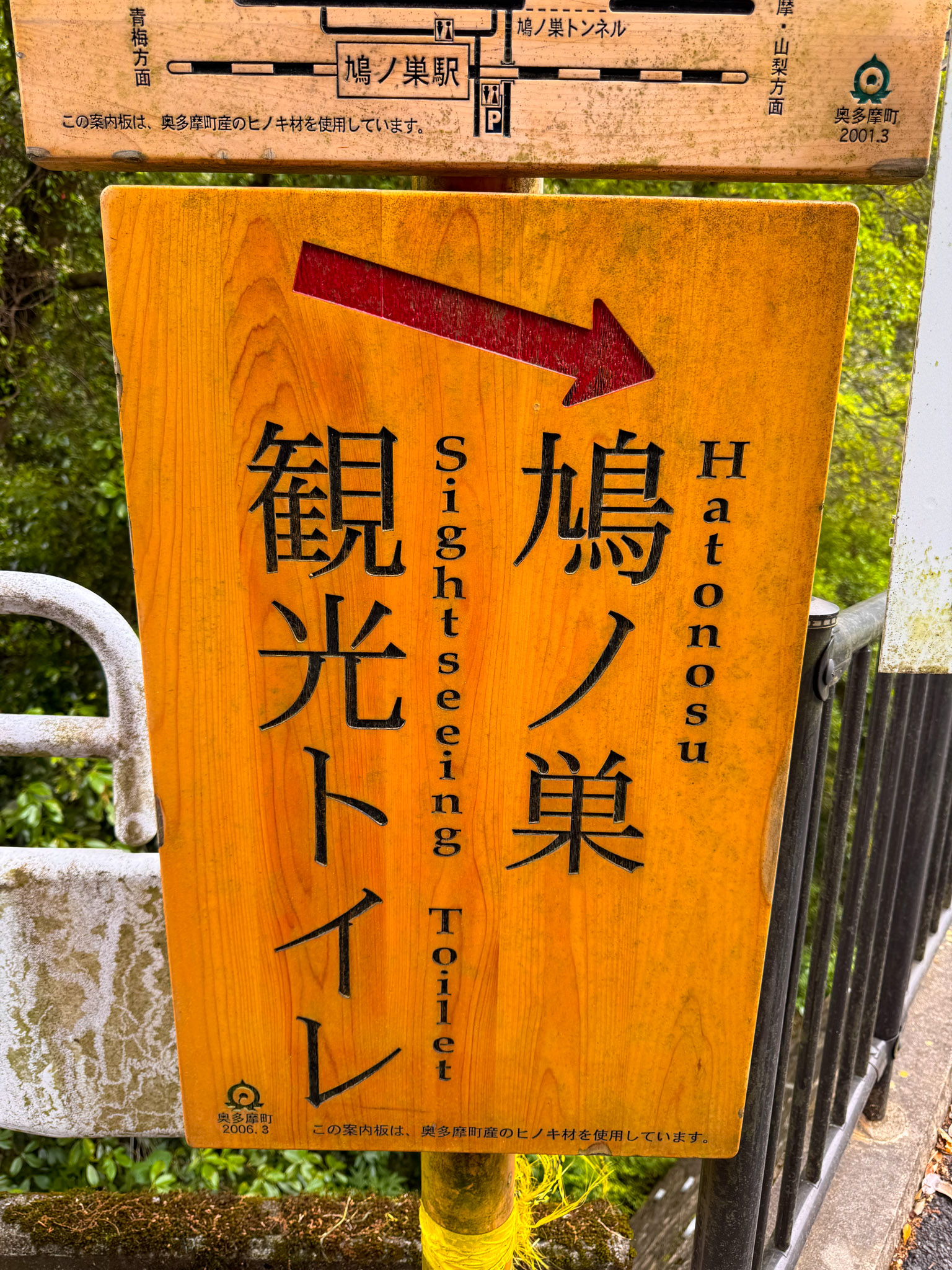 A carved wooden sign with a red arrow reading 鳩ノ巣 観光トイレ in Japanese and Hatonosu Sightseeing Toilet in English, mounted on a guardrail above the gorge