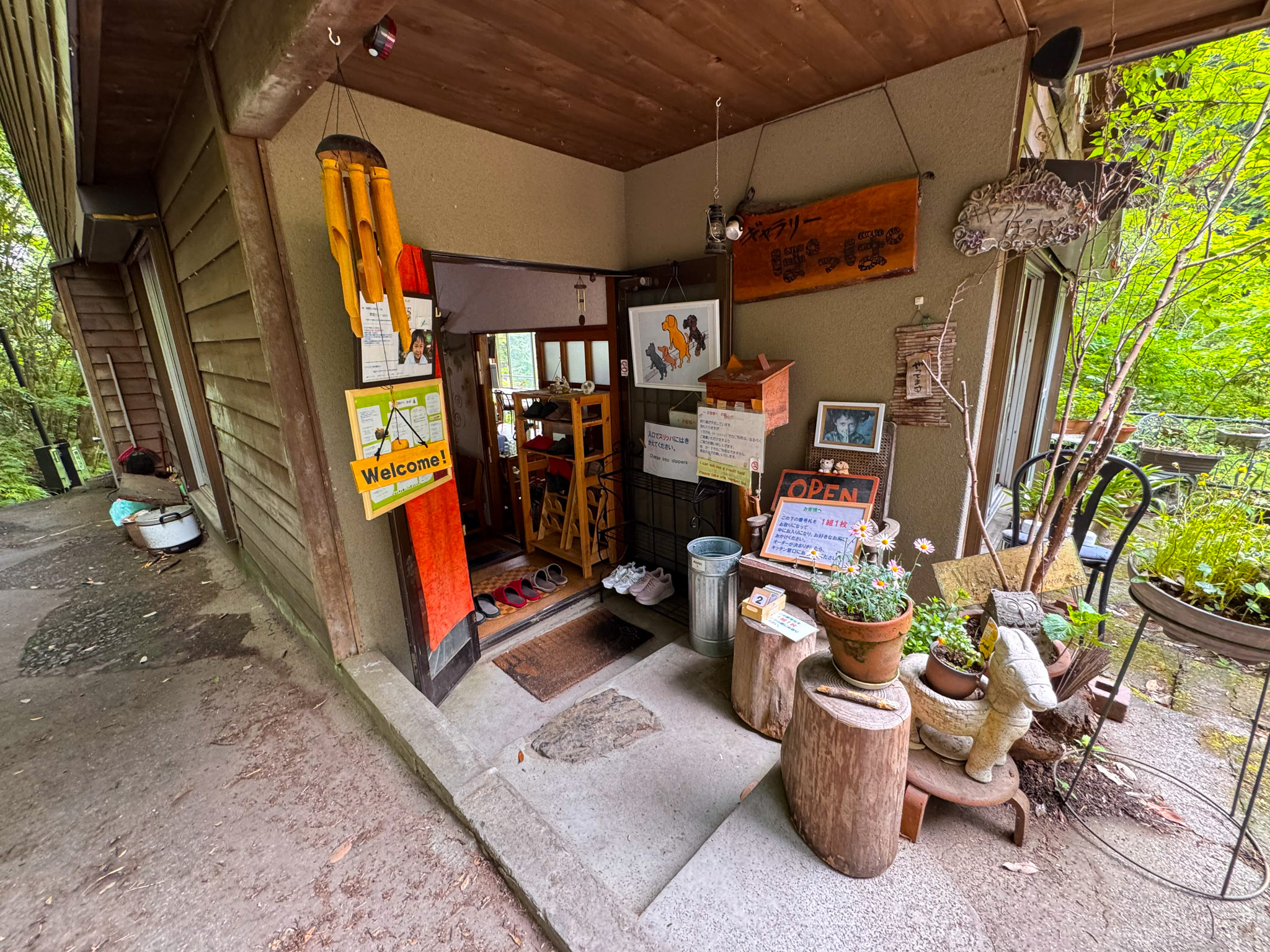 The entrance to Café Poppo — a small wooden cabin doorway with a Welcome sign, an OPEN board, potted plants, wind chimes, and an orange noren curtain