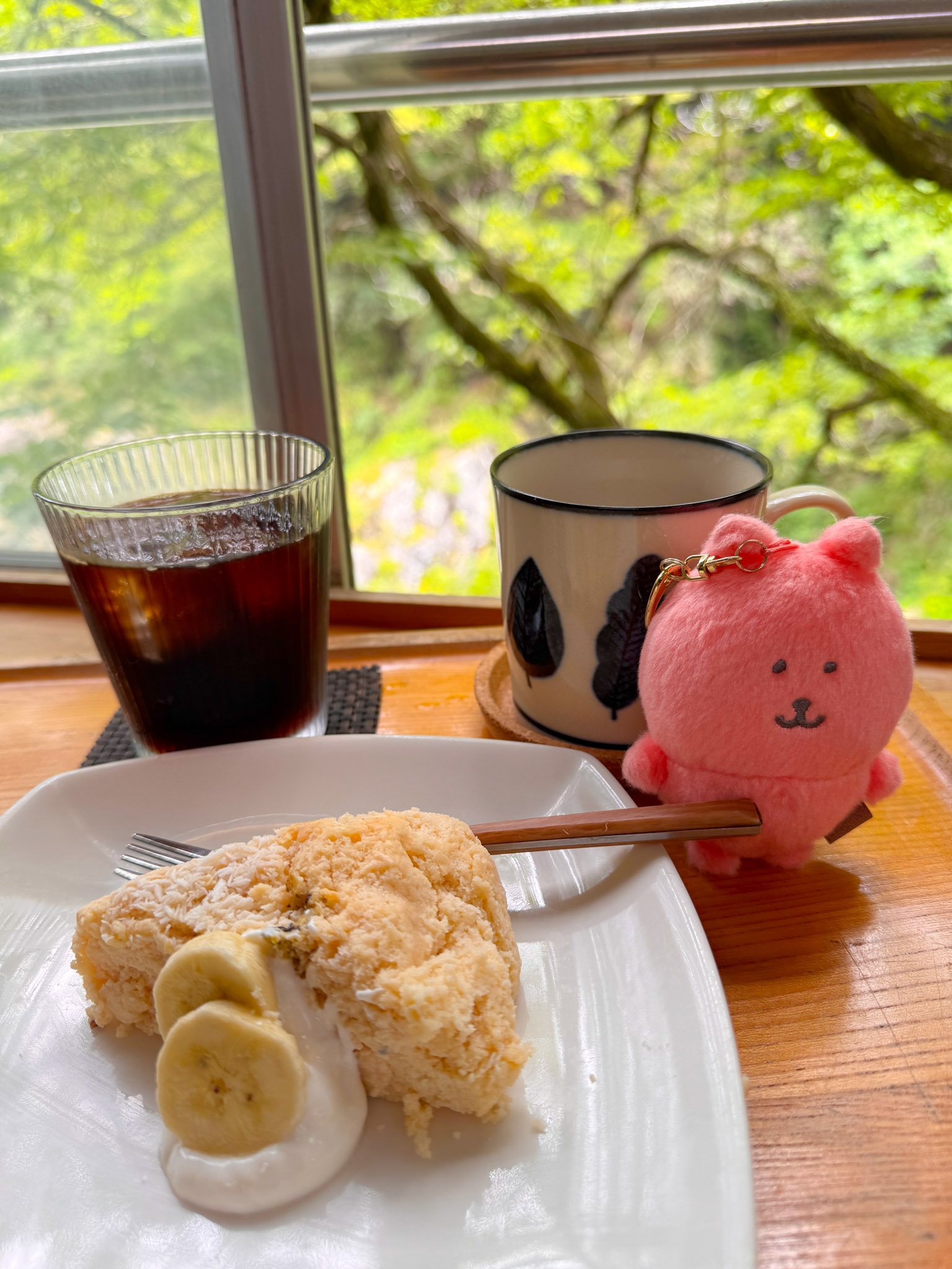A café table by the window — a slice of tofu cheesecake with banana and cream on a white plate, a glass of cold brew coffee, a leaf-patterned mug, and a small pink Joke Bear plush, all backlit by green forest outside