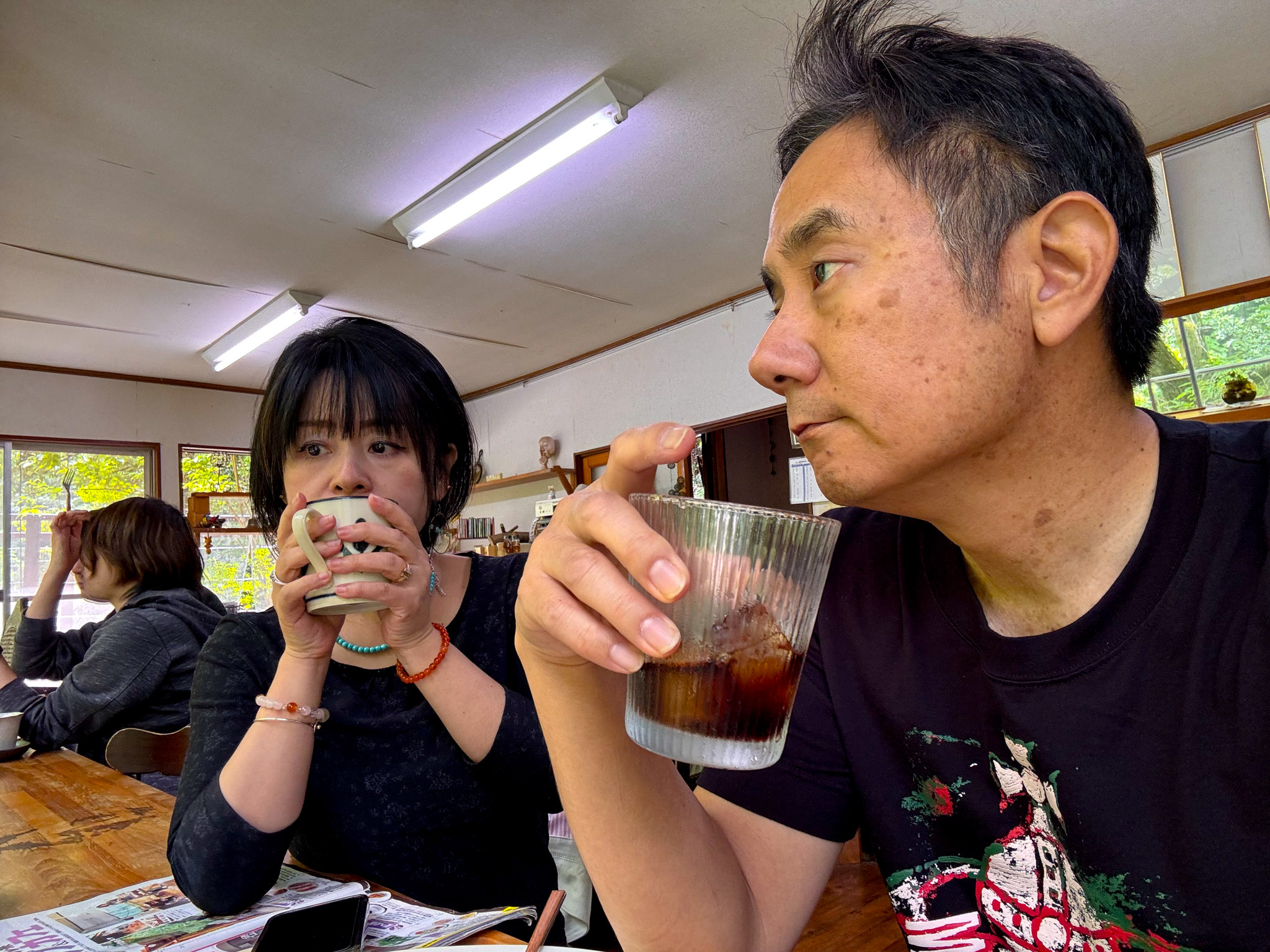 John and Ayumi sitting at the café table — John sipping iced coffee from a glass, Ayumi drinking from a mug, the bright forest visible through the window behind them