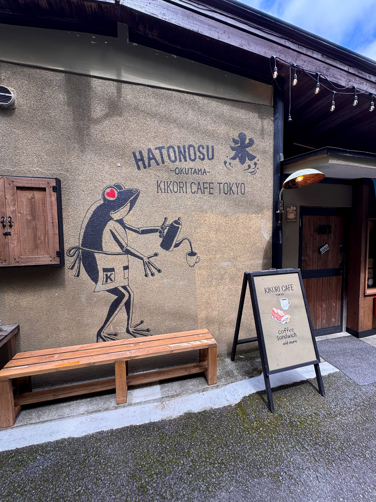 The exterior wall of Kikori Café Tokyo in Hatonosu — a large painted mural of a red-eyed frog pouring tea, with HATONOSU OKUTAMA and KIKORI CAFE TOKYO lettering and a sandwich-board menu out front