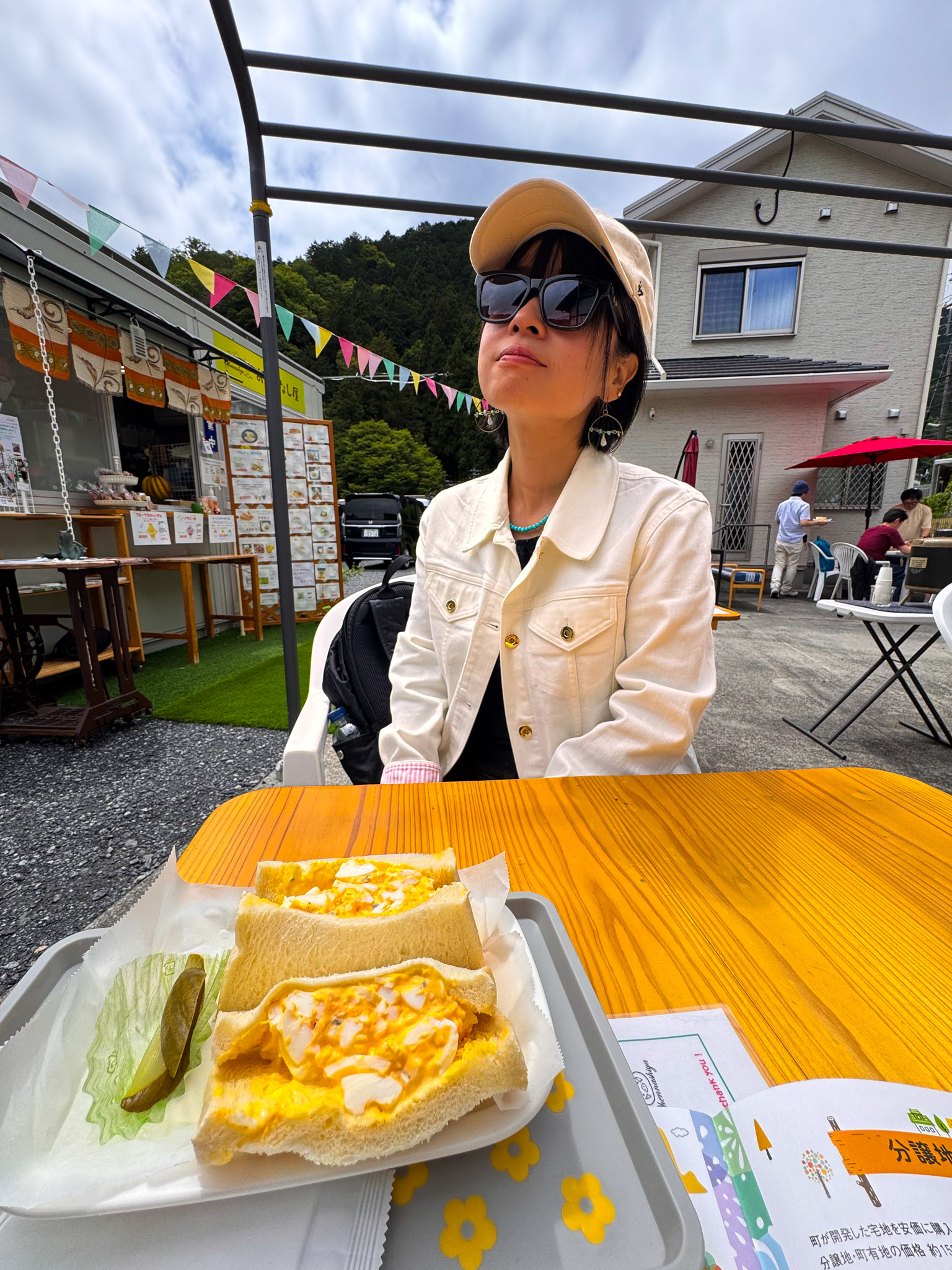 Ayumi in a cream jacket and sunglasses tilting her head back at an outdoor table, a tamago sando — egg salad sandwich with a pickle — on a tray in front of her