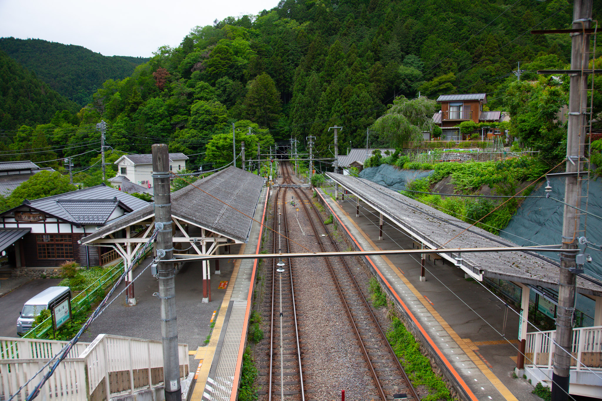 Hatonosu Station seen from the overhead footbridge — two rail tracks running between a traditional tiled-roof station building and tree-covered mountains, a small village tucked into the green hills