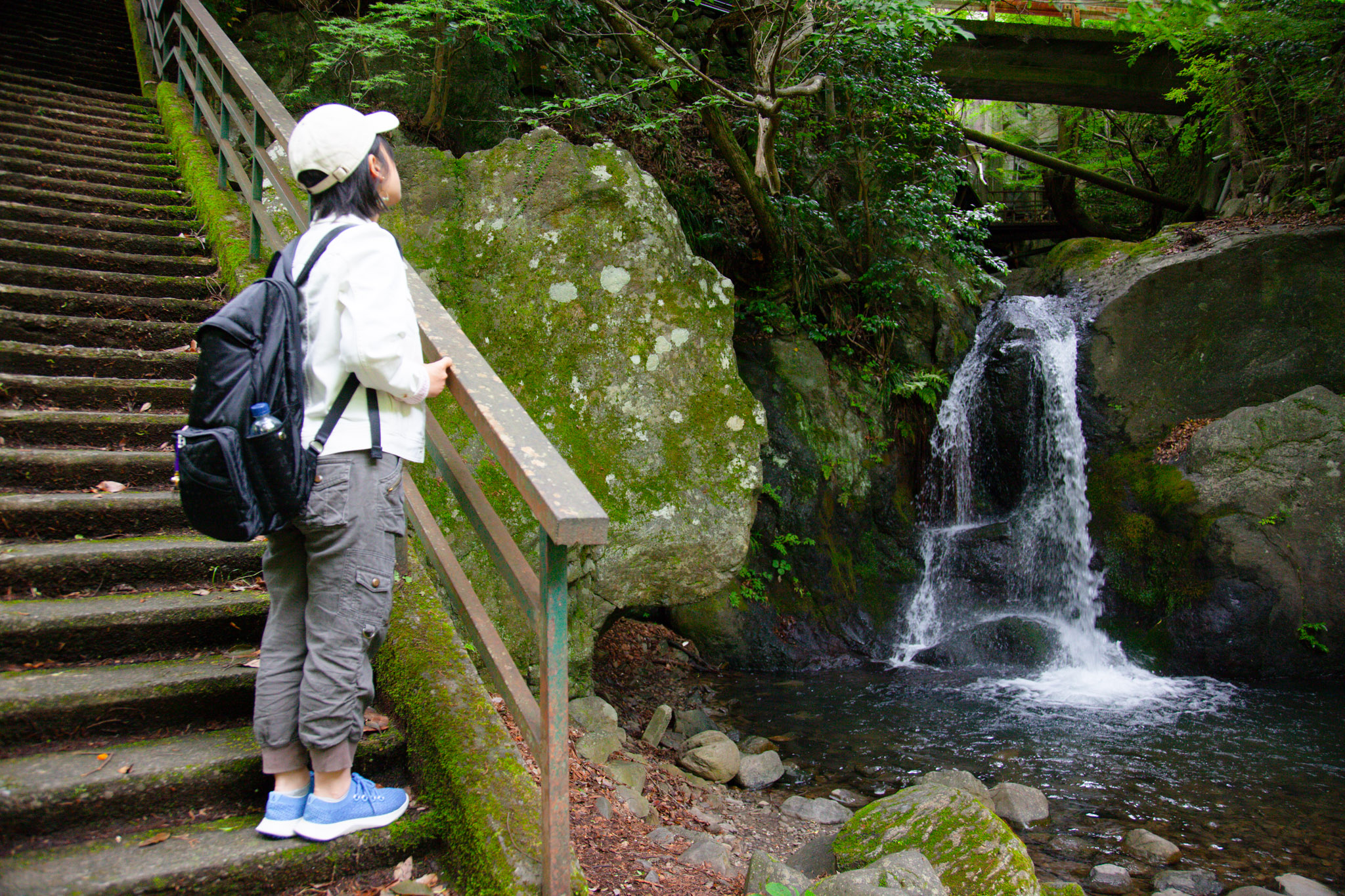 Ayumi in a white jacket and backpack pausing at a wooden handrail beside a small waterfall spilling over dark rock into a shaded pool