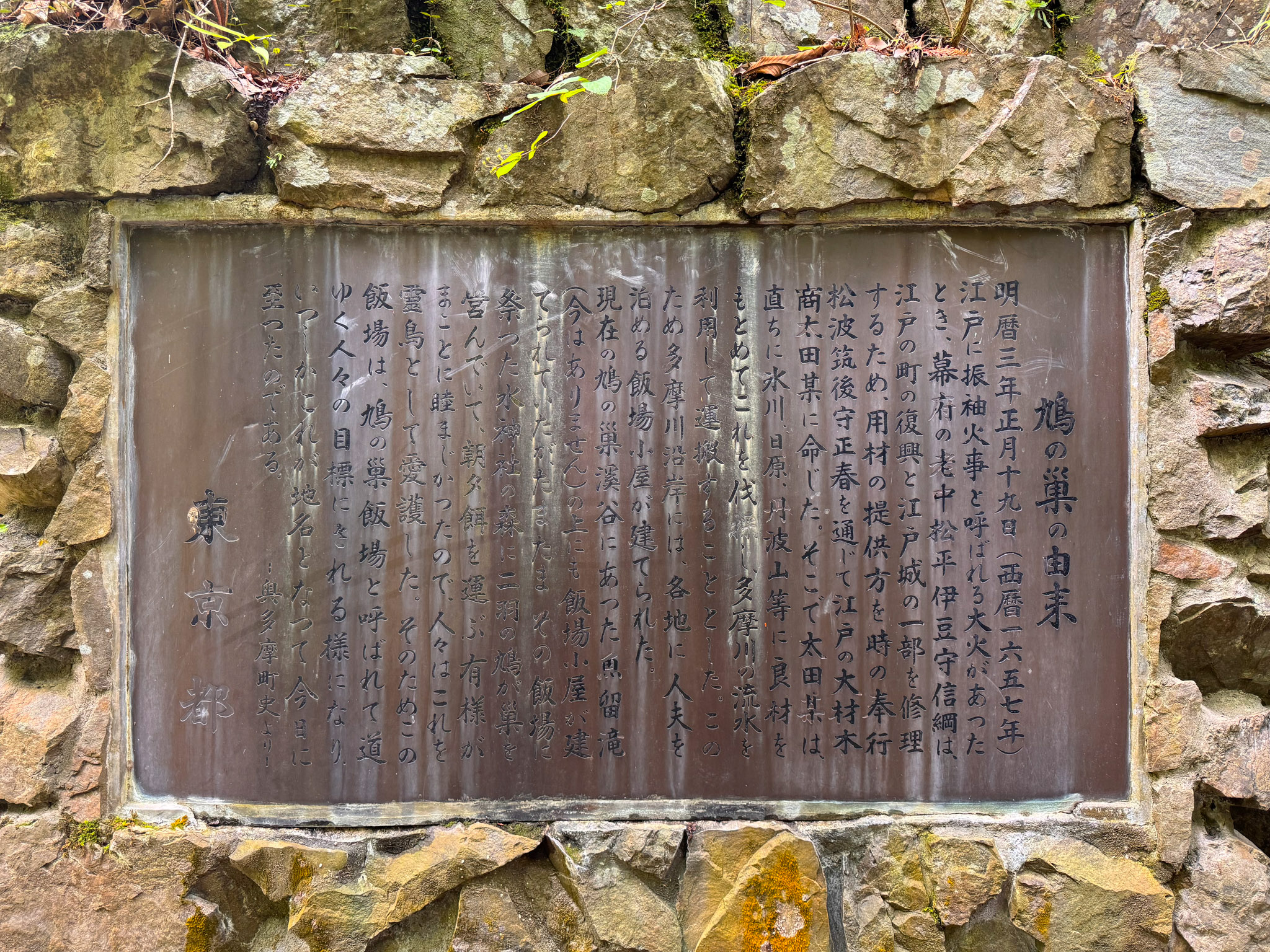 A weathered bronze plaque set into a stone wall, titled 鳩の巣の由来 (The Origin of Hatonosu), with several columns of Japanese text explaining the place name and signed 東京都 (Tokyo Metropolis)