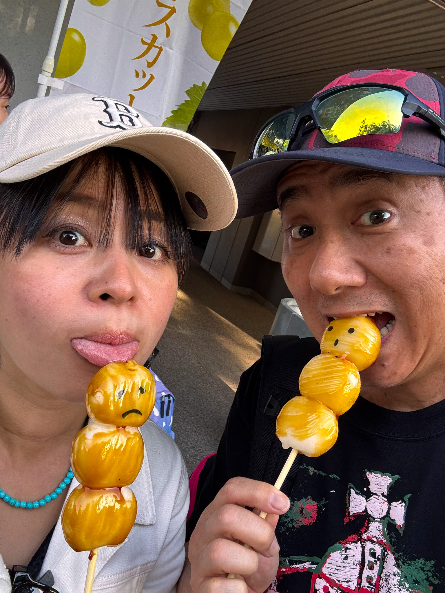 John and Ayumi taking a selfie, each biting into a skewer of glazed grilled mochi with painted faces — one smiling, one frowning — near a fruit shop in Kichijoji