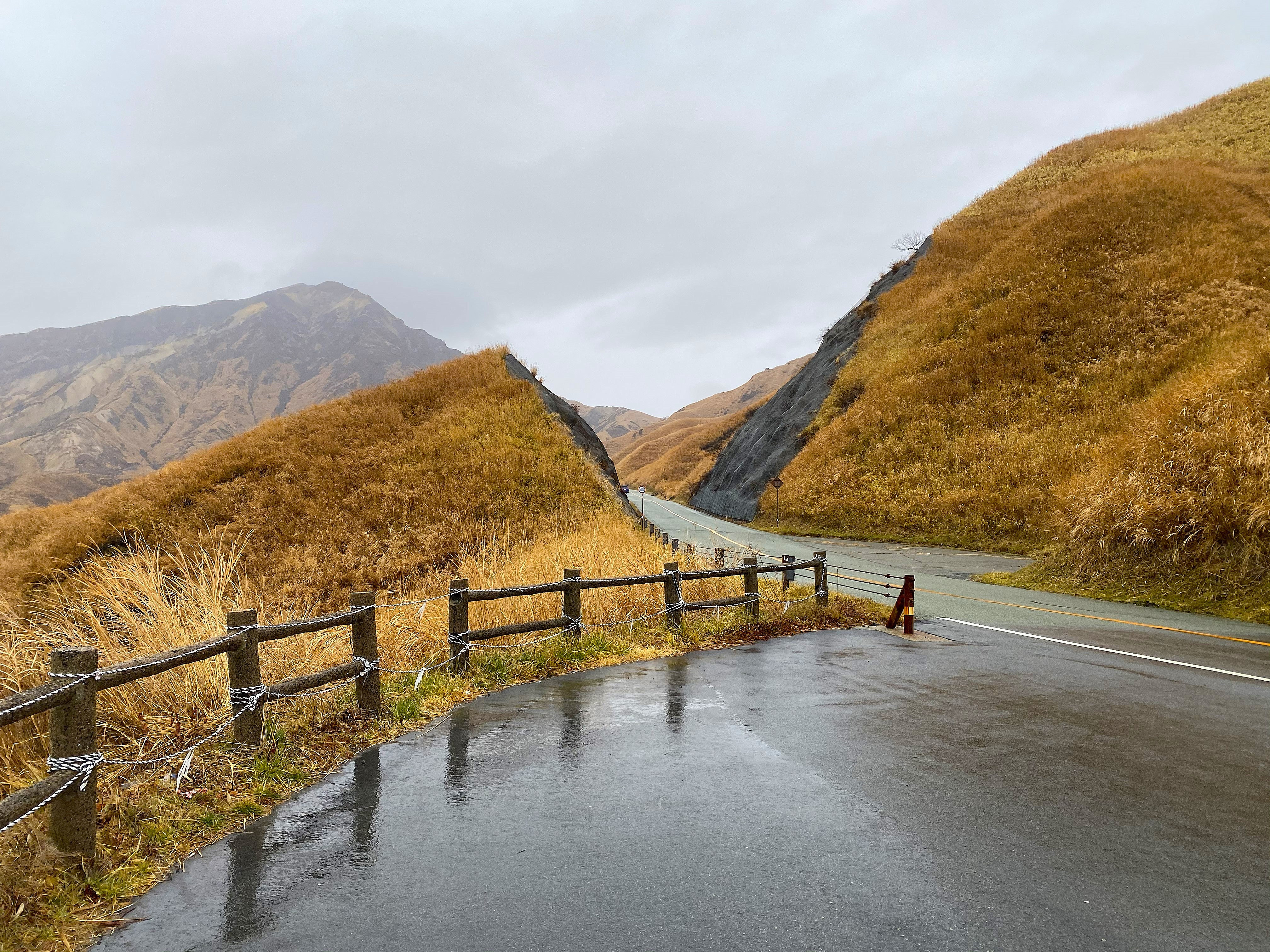 Wet mountain road through the golden grass hills of Aso heading toward Kurokawa — the road drops between two steep golden hillsides, wooden rope fence in the foreground, Aso mountains in the distance under overcast sky