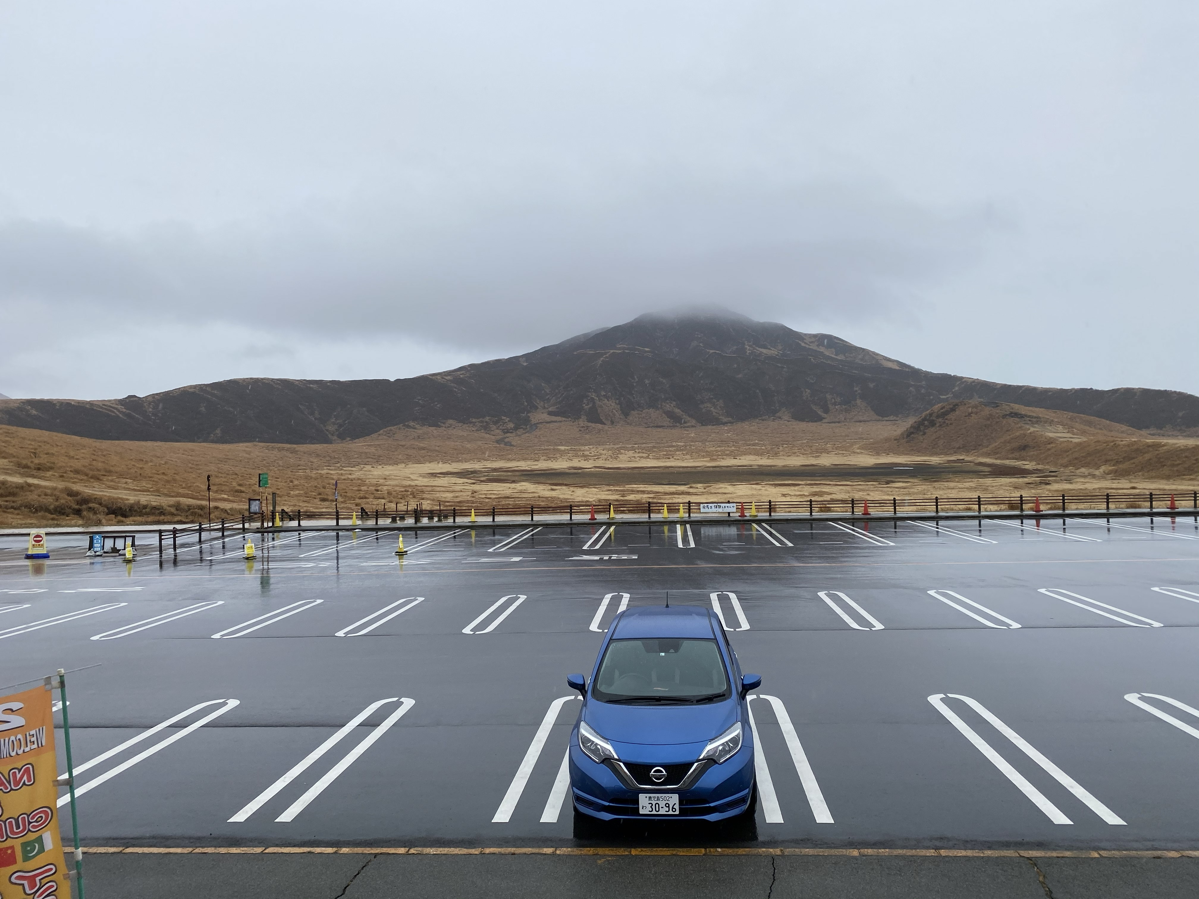 Blue Nissan Note rental car parked alone in a large wet parking lot near Mt. Aso — Mt. Eboshi looming directly behind, caldera grassland visible between, summit in cloud