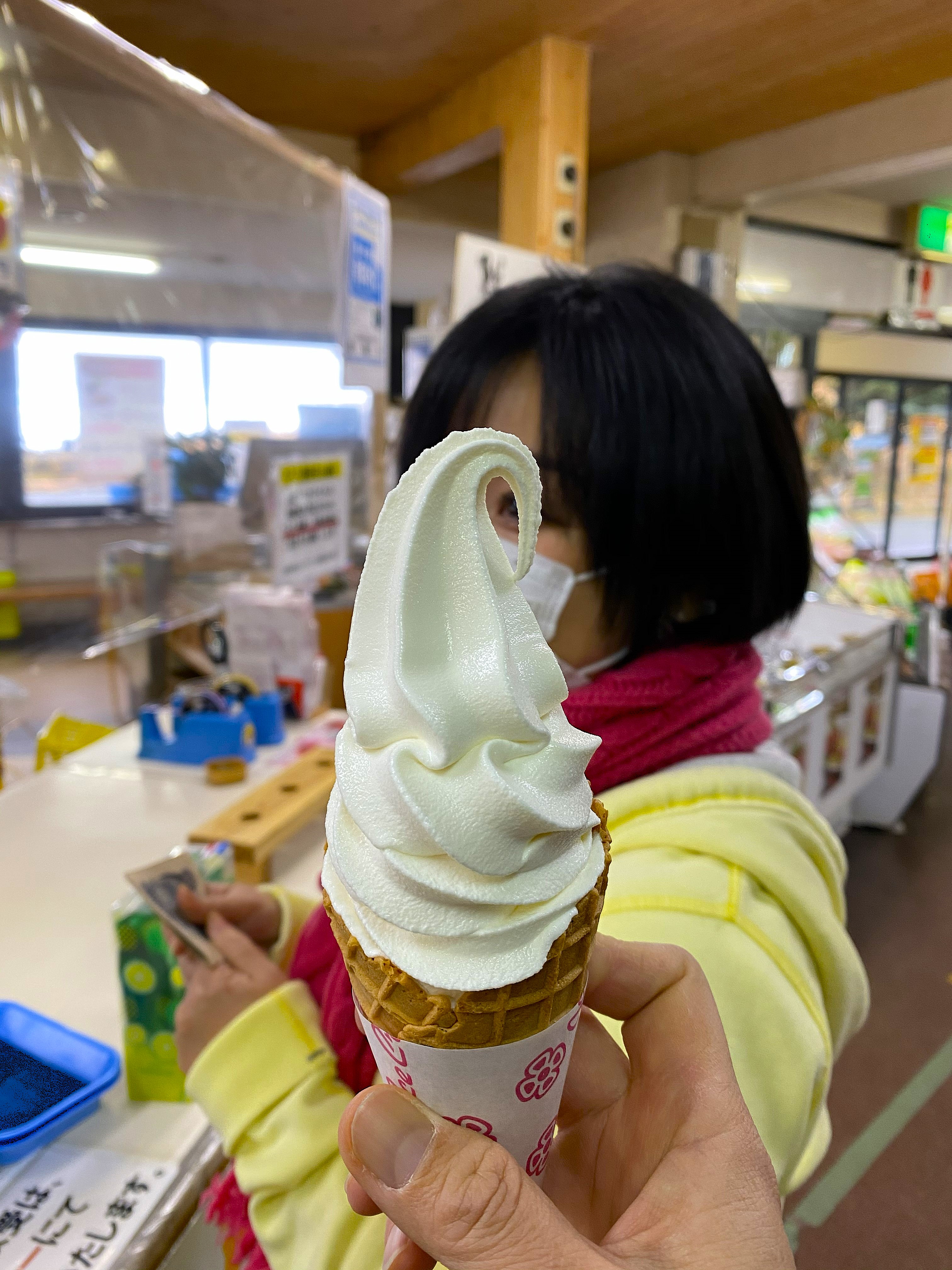 A tall, perfectly swirled Jersey milk soft serve ice cream cone held up at a roadside stop in the Aso region — Ayumi visible behind it in a yellow jacket and mask
