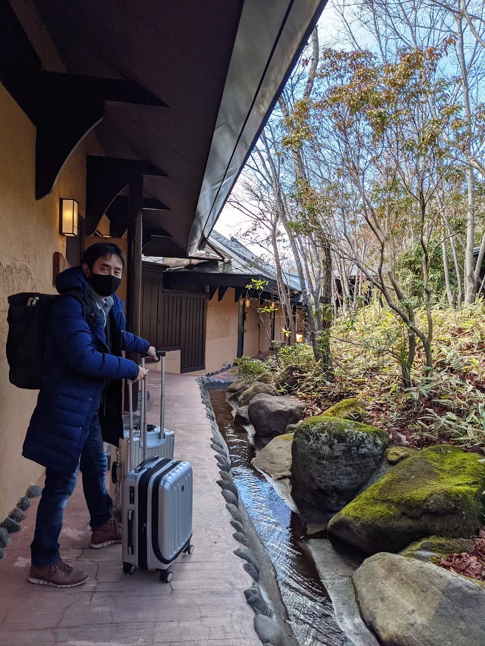 John standing with luggage on the covered entrance walkway at Satonoyu Waraku ryokan in Kurokawa Onsen — a small stream runs alongside the stone path, traditional Japanese architecture with dark timber and earth-toned plaster walls, bare winter trees beyond