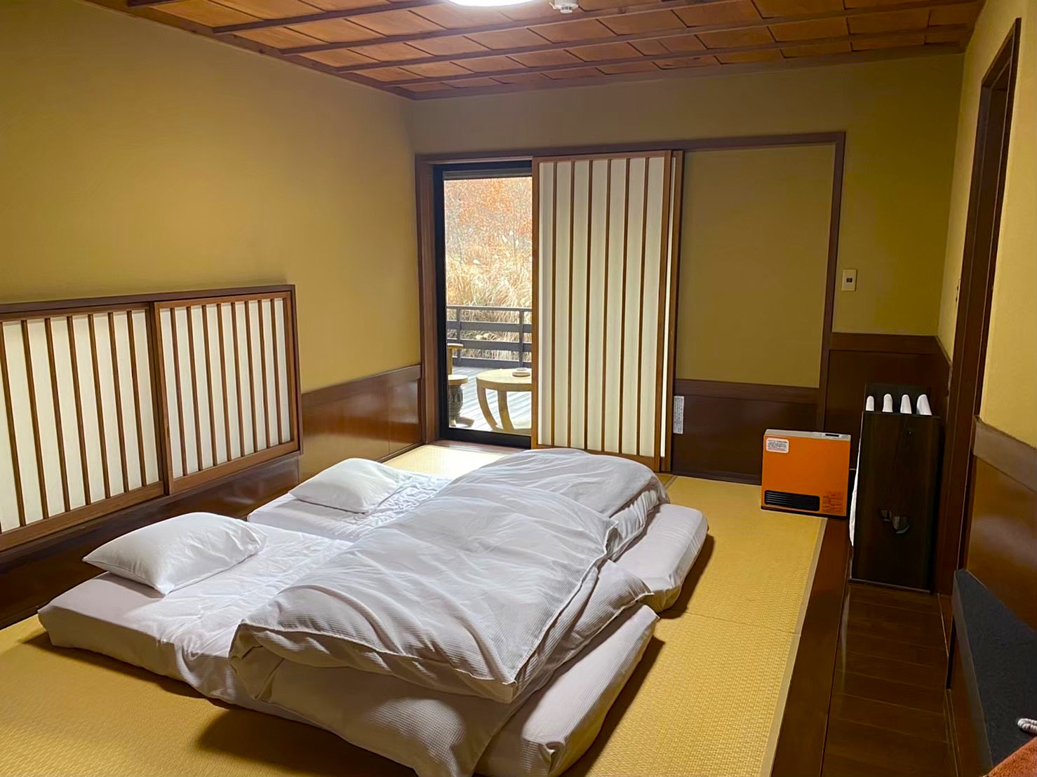 The tatami room at Satonoyu Waraku ryokan — futon laid out on the straw mat floor, sliding shoji screens open to an engawa deck with bare winter trees visible beyond, an orange kerosene heater in the corner