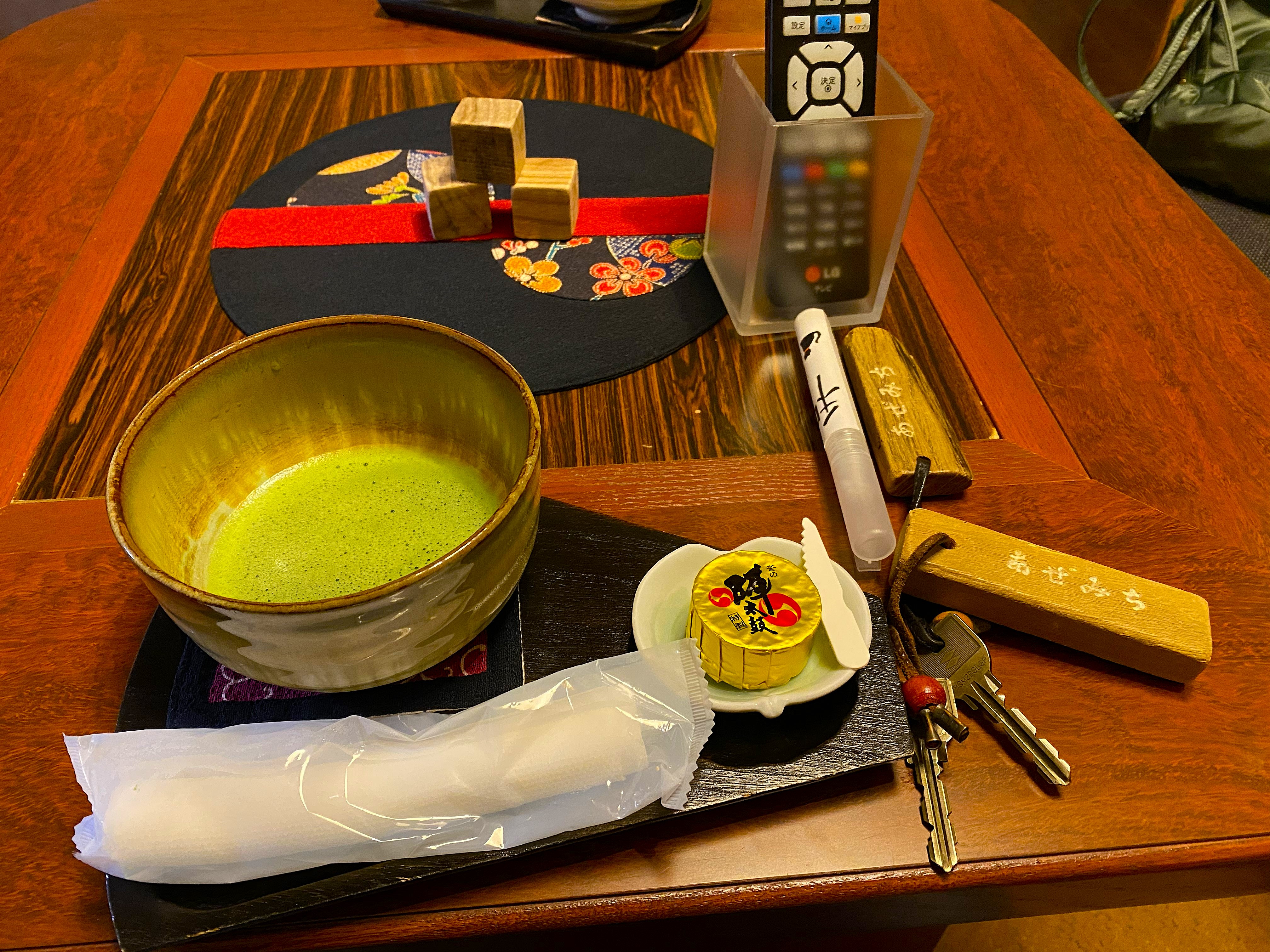 Welcome matcha in a handmade ceramic bowl on a dark lacquered tray, beside a wrapped wagashi sweet, and the room key with a wooden fob inscribed あぜみち — the room name Azemichi