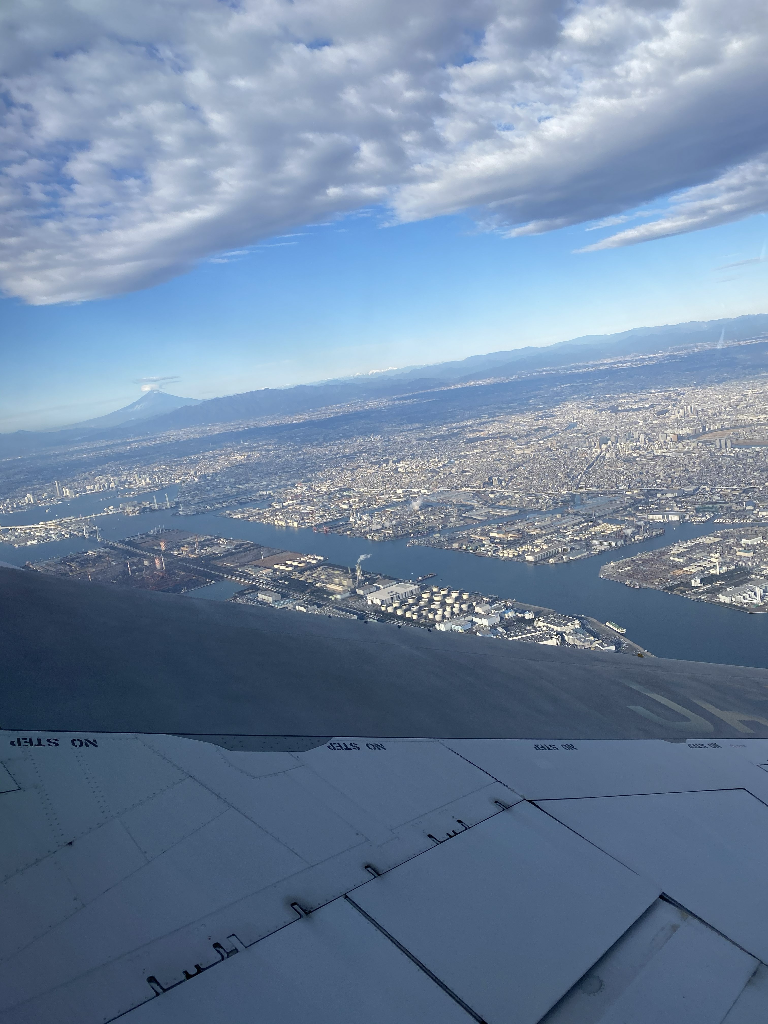 Aerial view from the plane window departing Haneda — Tokyo Bay and industrial waterways below the wing, Mount Fuji faintly visible on the far horizon beneath a bank of clouds
