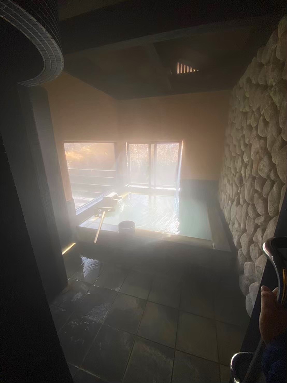 The private onsen bath at Satonoyu Waraku ryokan — milky hot spring water steaming in a stone-walled room, wooden ladle resting on the edge, windows overlooking bare winter trees, late afternoon light filtering through the steam