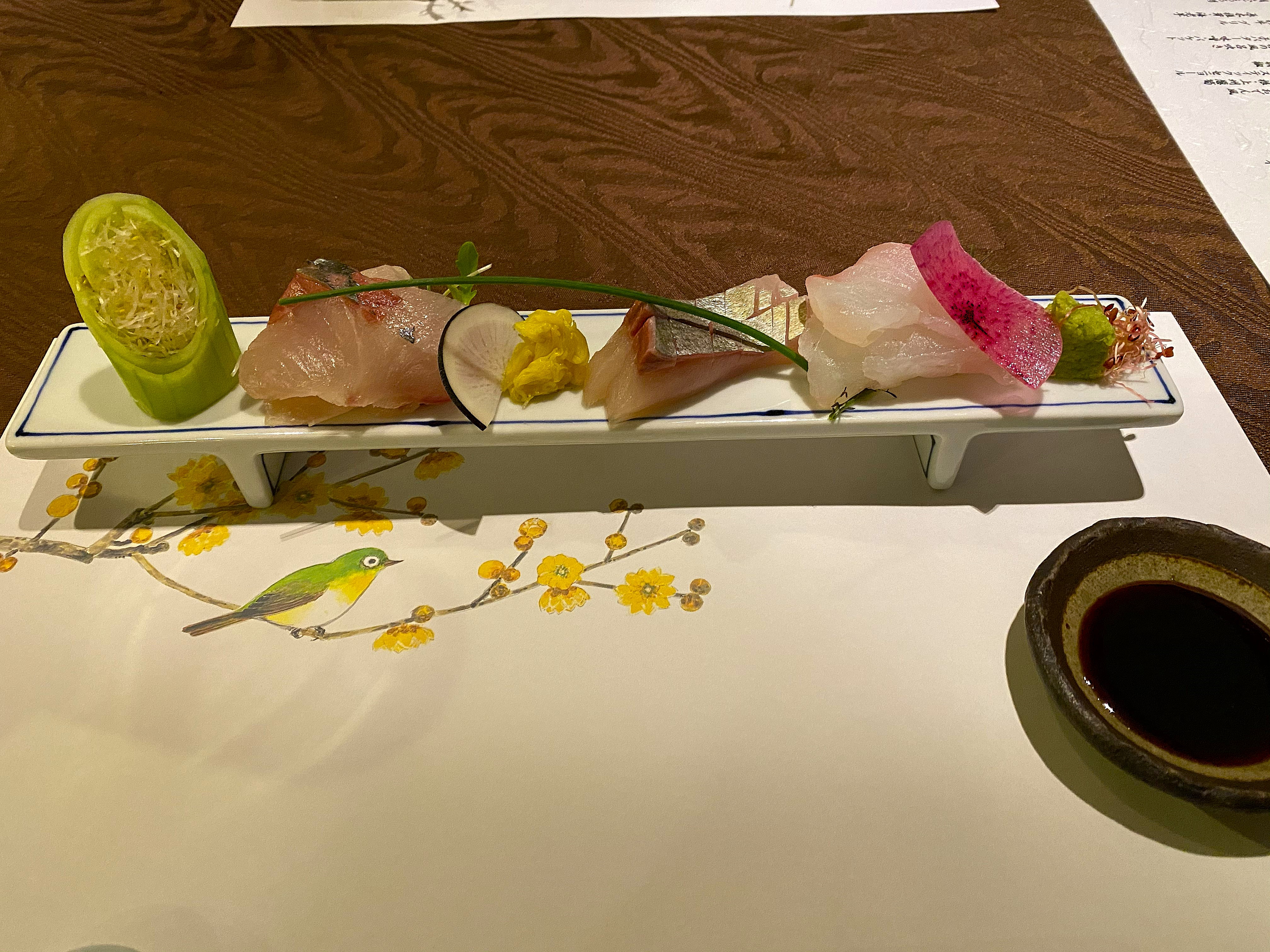 Sashimi course at Satonoyu Waraku — pieces of sea bass, octopus, and grouper arranged on a long narrow white plate painted with a mejiro bird on plum blossoms, daikon julienne in a cucumber cylinder, soy sauce in a small ceramic vessel