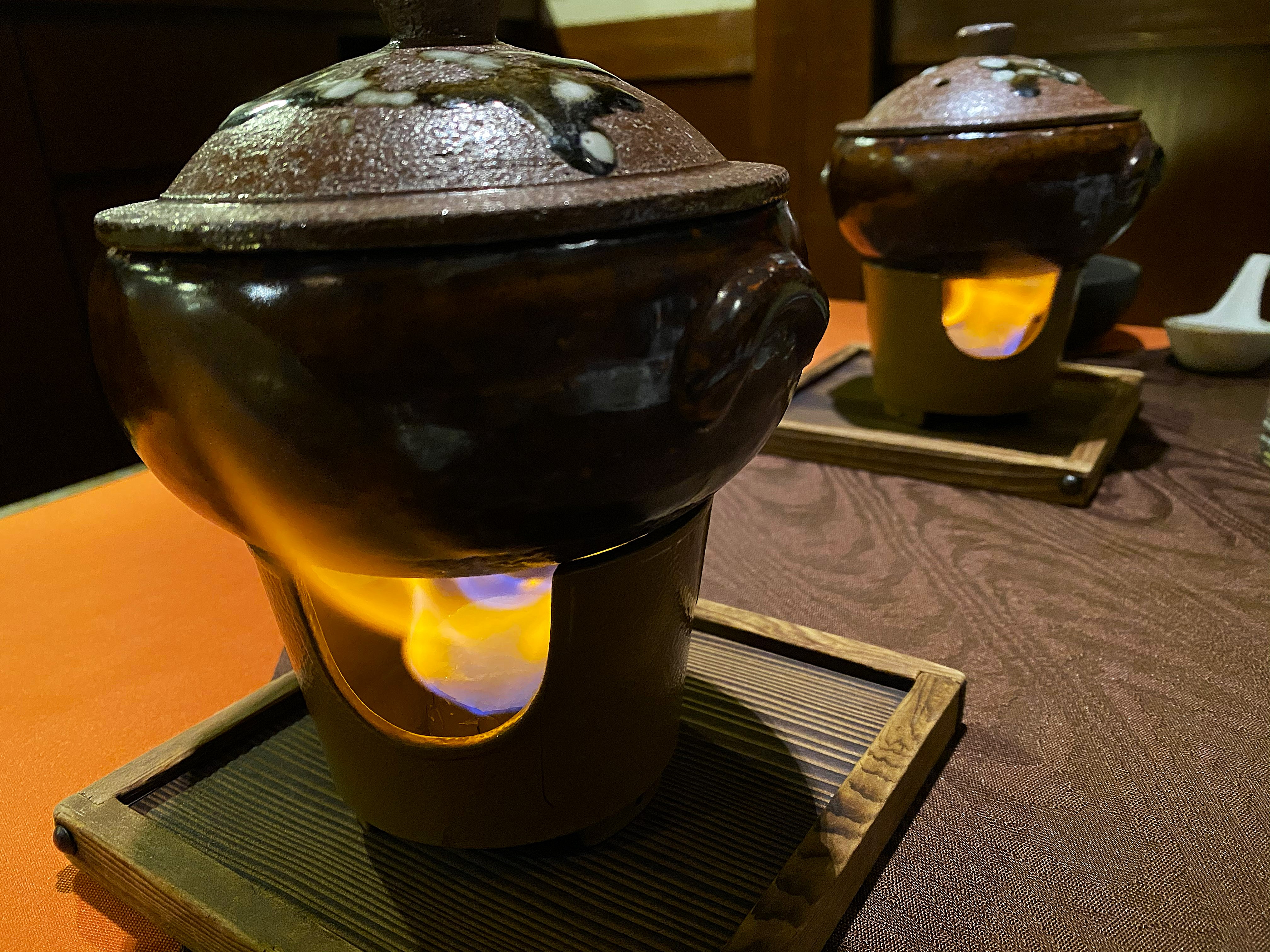 Two dark glazed donabe earthenware pots on individual wooden-framed alcohol burners at the kaiseki dinner table — flames visible through the ceramic bases, both pots steaming with lids on