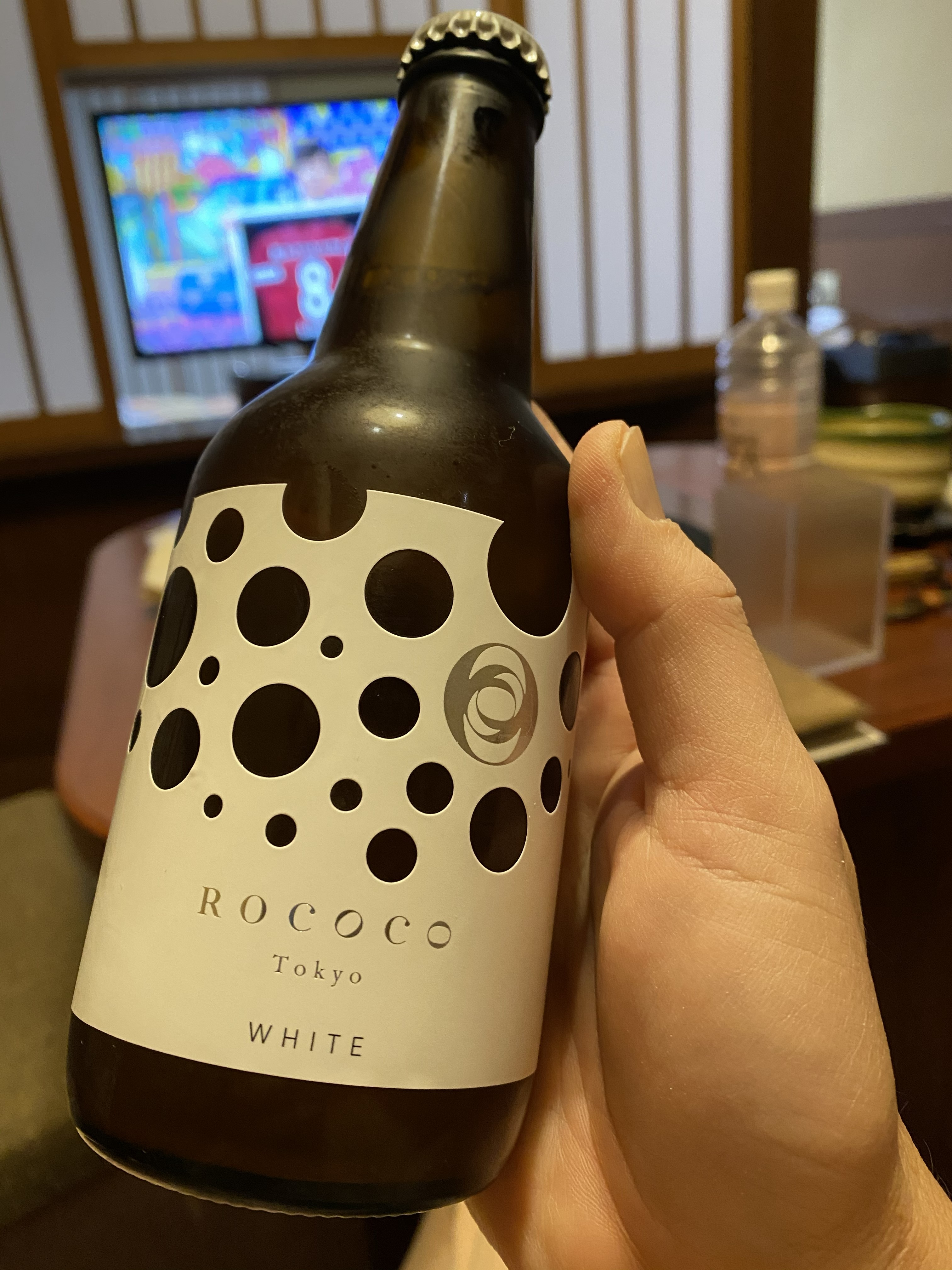 A bottle of ROCOCO Tokyo White craft drink held up in the ryokan room — the distinctive polka-dot label against a dark amber bottle, Japanese TV playing in the background
