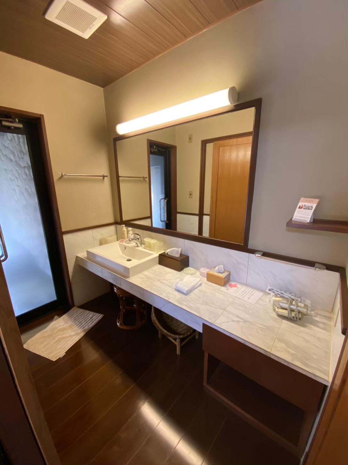 The dressing and vanity area of the private bath at Satonoyu Waraku — marble counter with a square vessel sink, wide mirror with warm lighting, cedar ceiling, toiletries and yukata essentials laid out on the counter