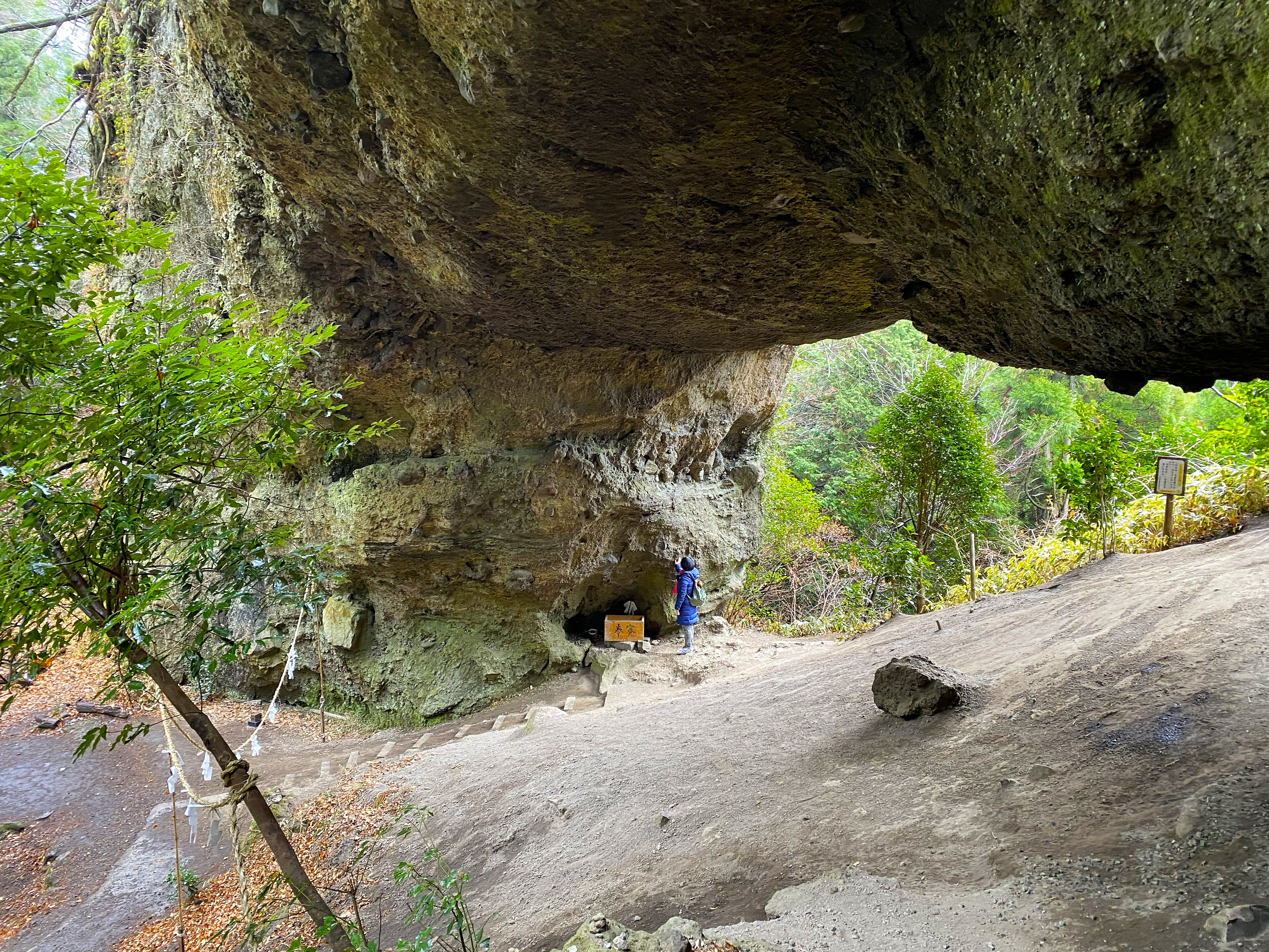 Inside the massive volcanic rock overhang at Kamishikimi Kumanoimasu Shrine — Ayumi photographs the small cave shrine tucked into the cliff face, shimenawa rope in the foreground, the arch framing green forest beyond