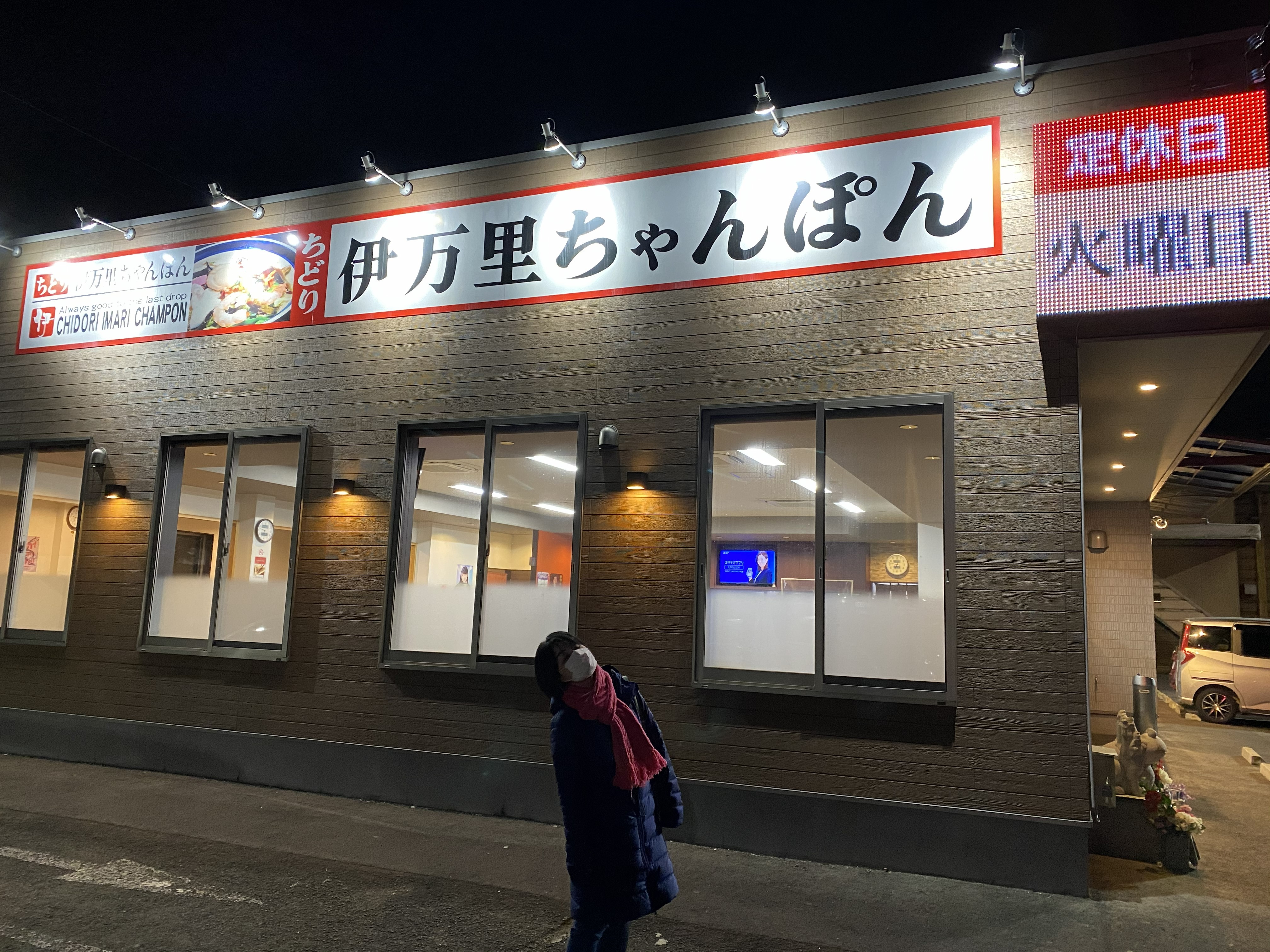 Chidori Imari Champon restaurant exterior at night — the large red-bordered sign reading 伊万里ちゃんぽん brightly lit, Ayumi in a blue puffer jacket standing outside looking up at the sign