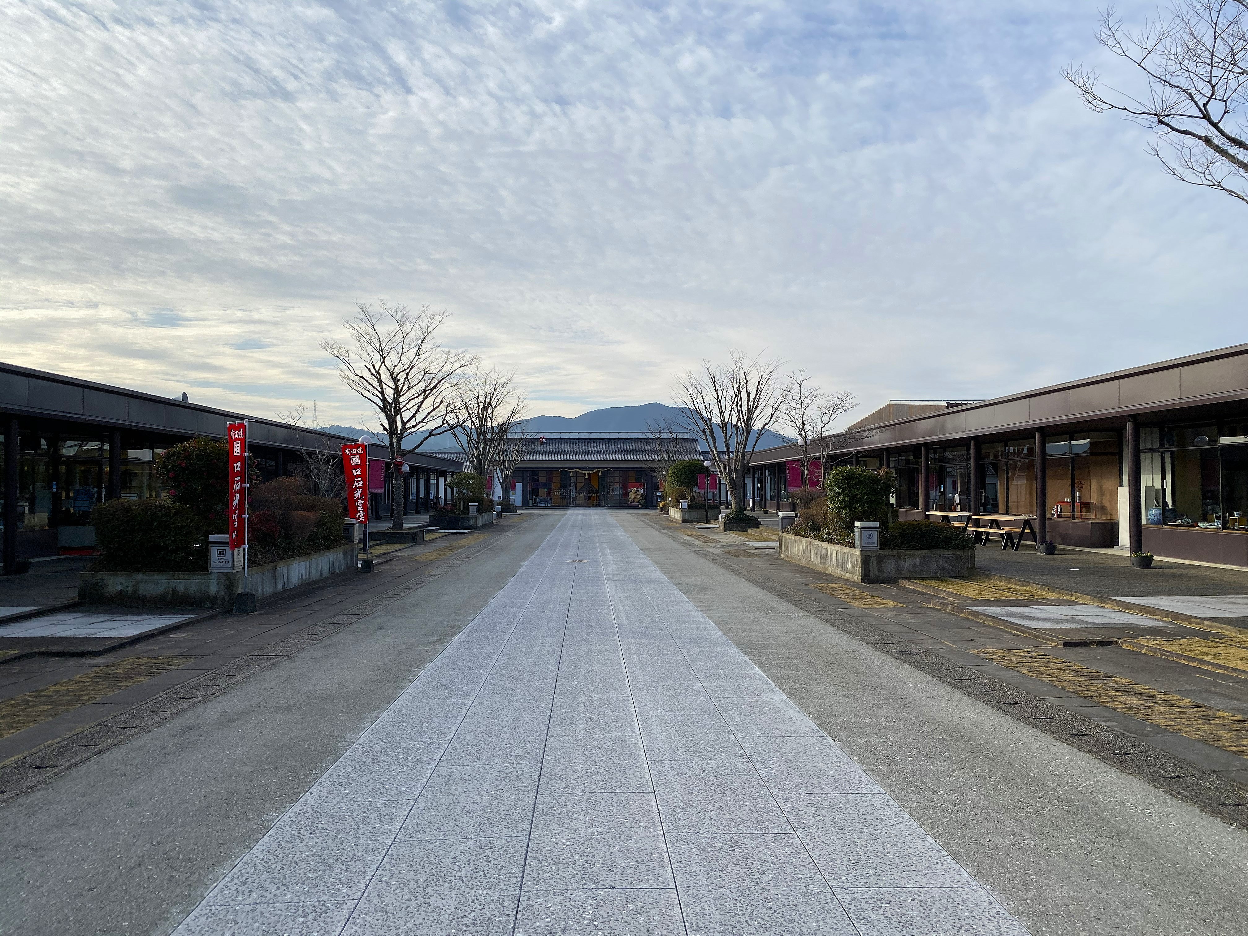 The main promenade at Arita Sera — a wide stone walkway stretching toward a traditional-style building at the far end, shops with dark timber facades on both sides, bare winter trees planted along the path, mountains visible in the background under a mackerel sky