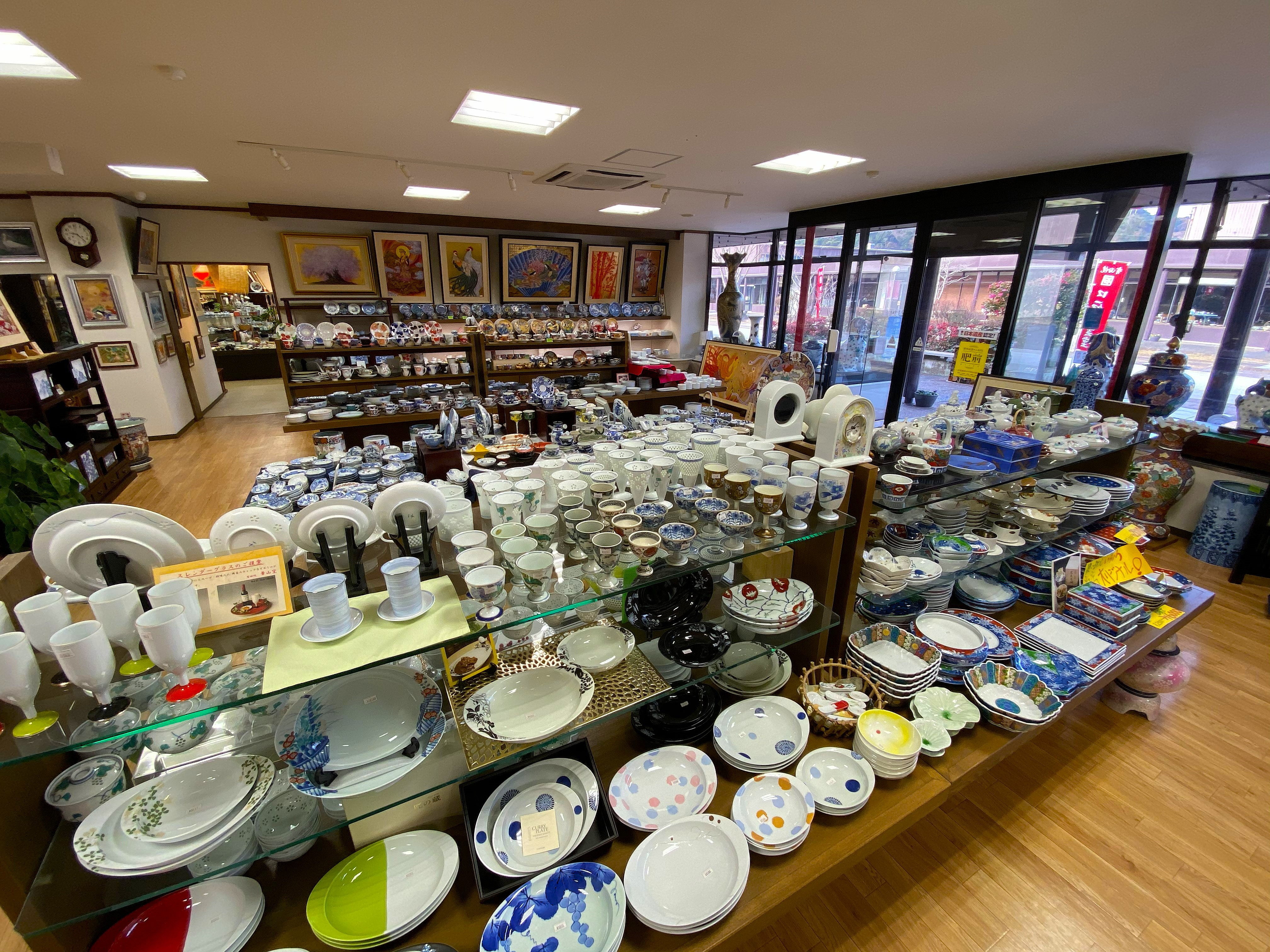 Inside one of the Arita Sera shops — wide-angle view of tables and shelves covered in porcelain pieces from multiple makers: plates, bowls, cups, vases in blue and white and colorful Imari styles, ukiyo-e style prints on the walls above