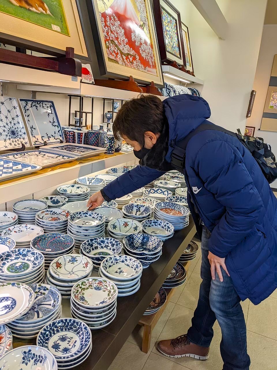 John leaning over a table inside an Arita Sera shop, examining stacks of blue and white hand-painted porcelain plates — various patterns in the classic Arita style, rows of plates of different sizes on shelves behind