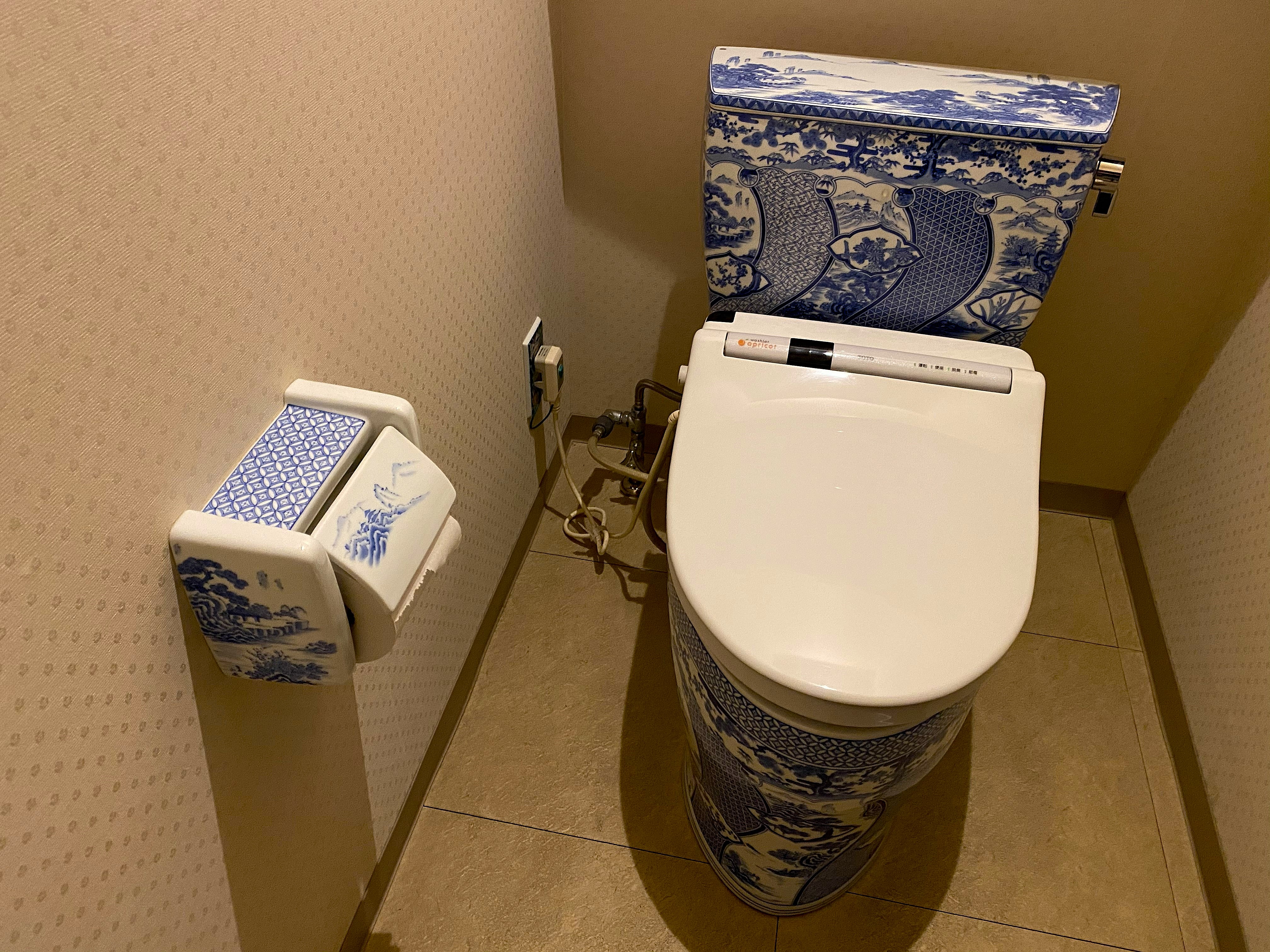 The Arita Sera porcelain toilet — blue and white hand-painted Arita porcelain covering the toilet tank and base, a traditional Japanese landscape scene painted on the ceramic, matching blue and white paper holder on the wall beside it