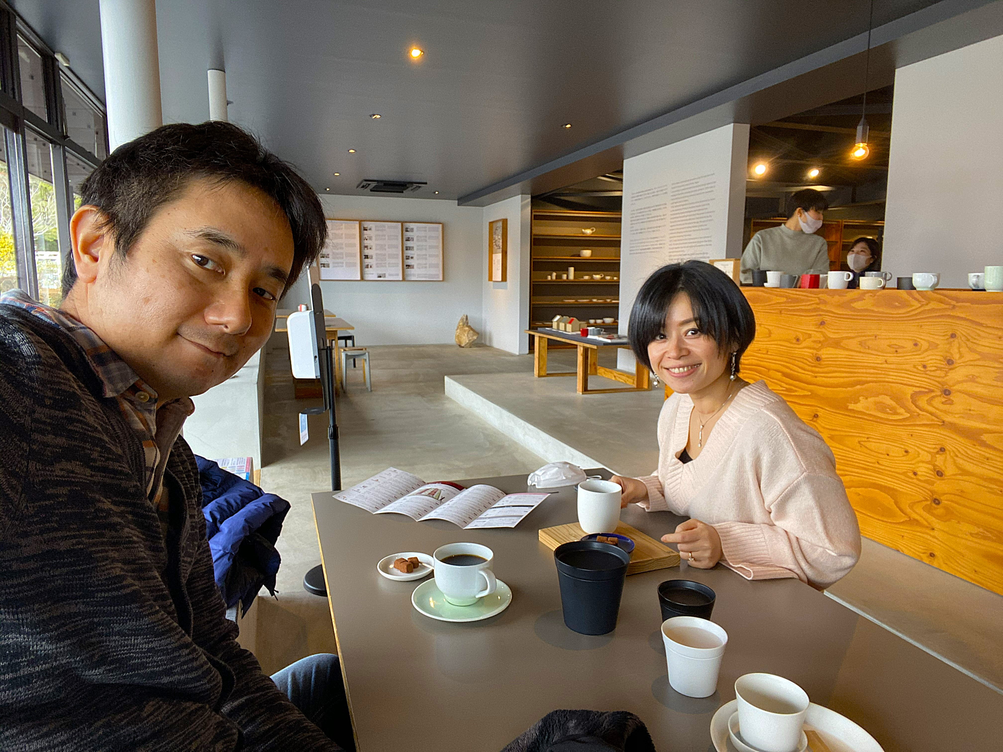John and Ayumi at a table inside a cafe at Arita Sera — both smiling at the camera with coffee and small sweets between them, the clean modern gallery-style interior visible behind, ceramic pieces displayed on shelves along the wall