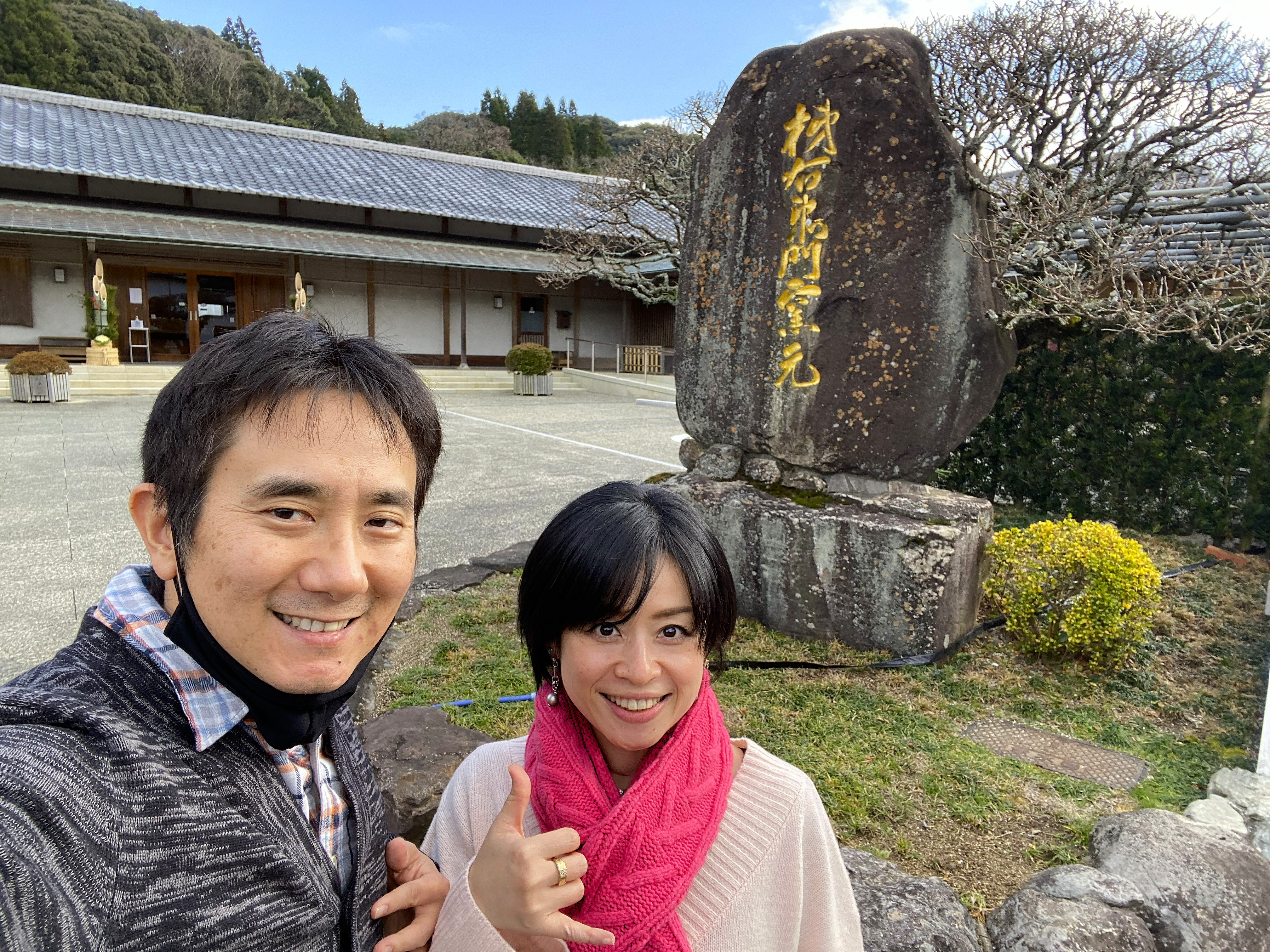 John and Ayumi selfie in front of the large stone at the Kakiemon kiln entrance — the stone reads 柿右衛門窯元 in gold characters, the traditional kiln buildings and bare trees visible behind them, blue sky above