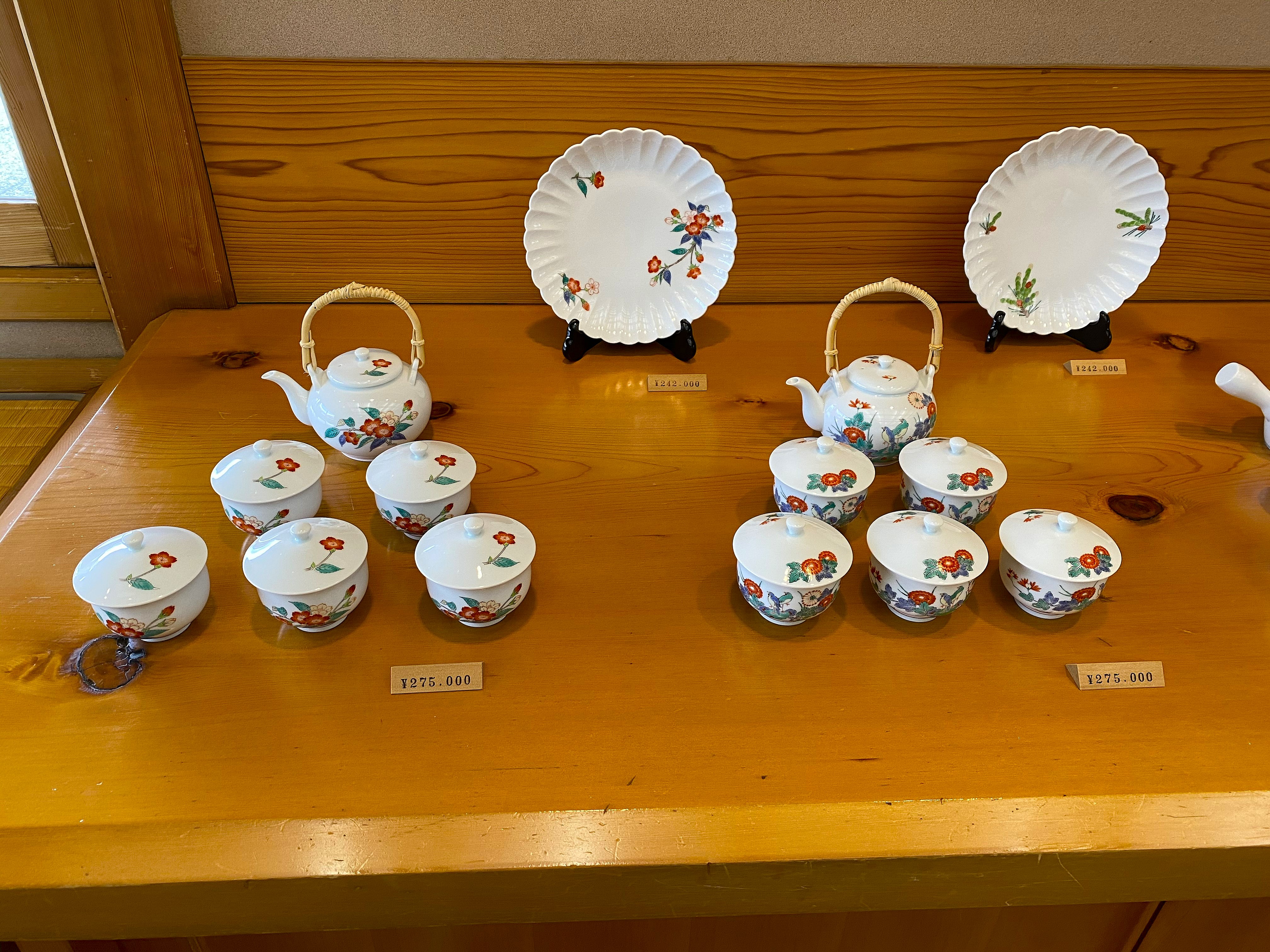 Kakiemon porcelain tea sets on display at the kiln showroom — two complete sets each with a bamboo-handled teapot and five lidded cups in the classic Kakiemon palette of red, blue and green flowers on warm white, priced at ¥275,000 per set; two large decorative plates on stands at ¥242,000 each