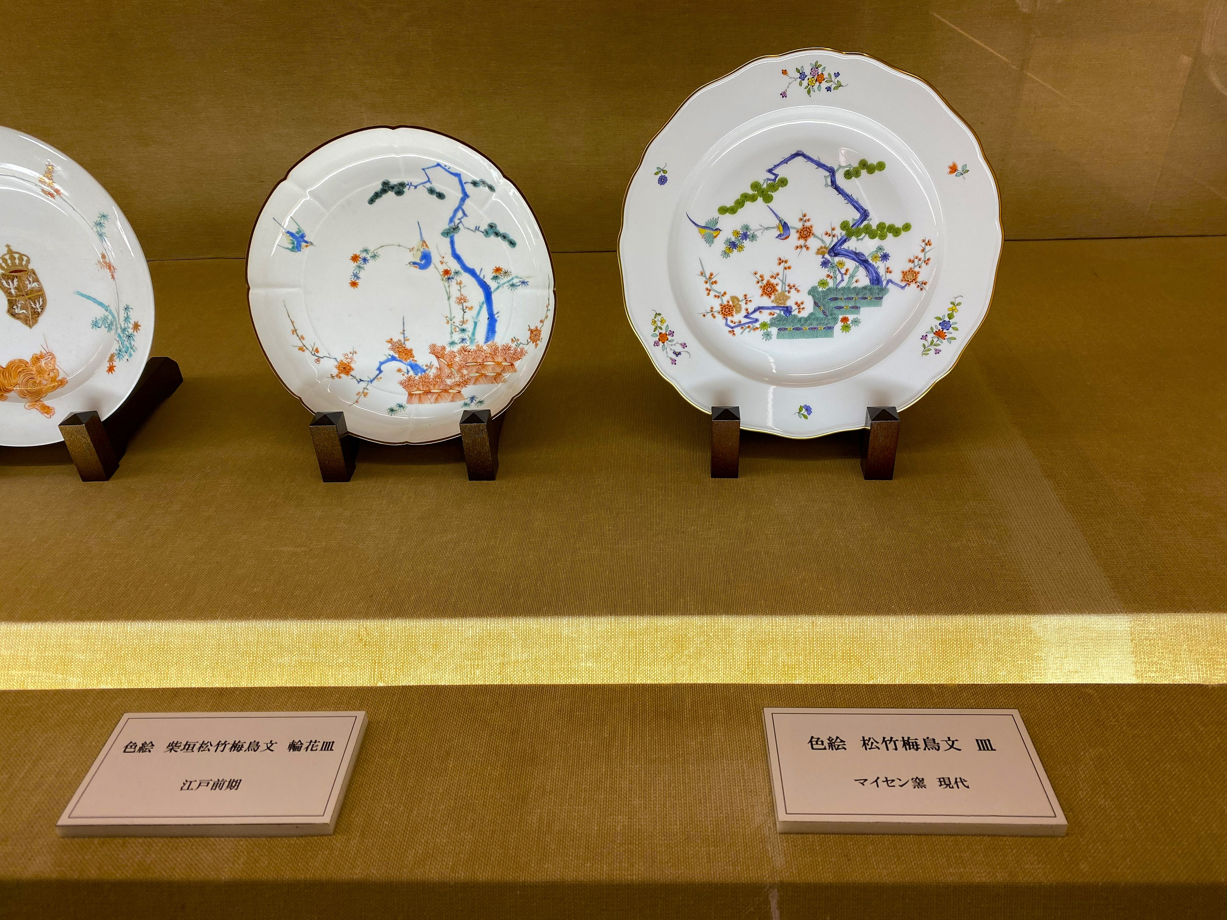 Display case at the Kakiemon kiln showing two plates side by side — left: 色絵 柴垣松竹梅鳥文 輪花皿, an Early Edo period Kakiemon plate with the pine-bamboo-plum-bird pattern; right: the same pattern reproduced on a modern Meissen plate — demonstrating the direct influence of Kakiemon on European porcelain