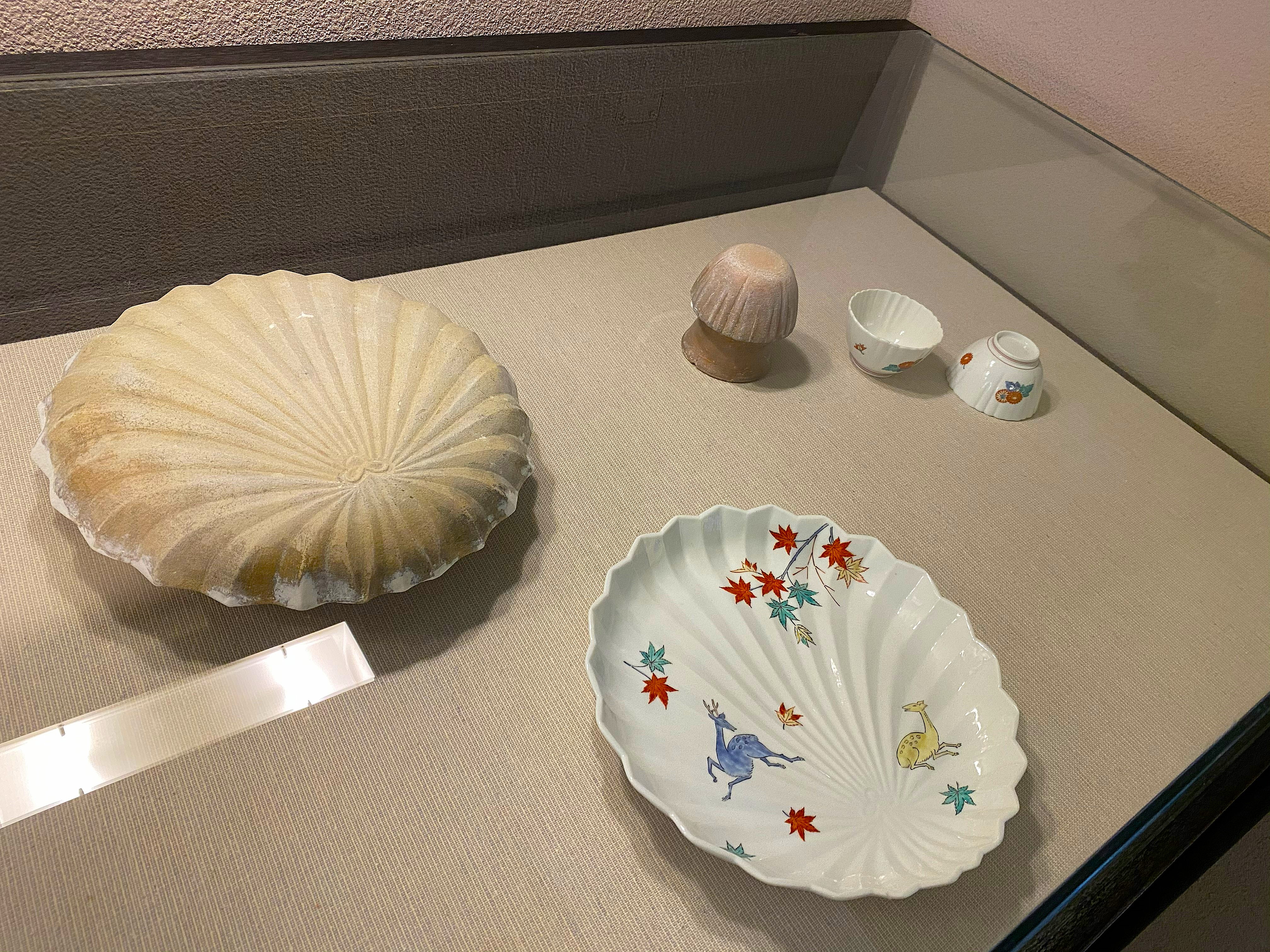 Display case showing Kakiemon production molds — a flat fan-shaped plate mold and a dome-shaped cup mold in unglazed gray clay, alongside a finished Kakiemon dish painted with deer and autumn maple leaves in the classic palette, and small finished Kakiemon cups