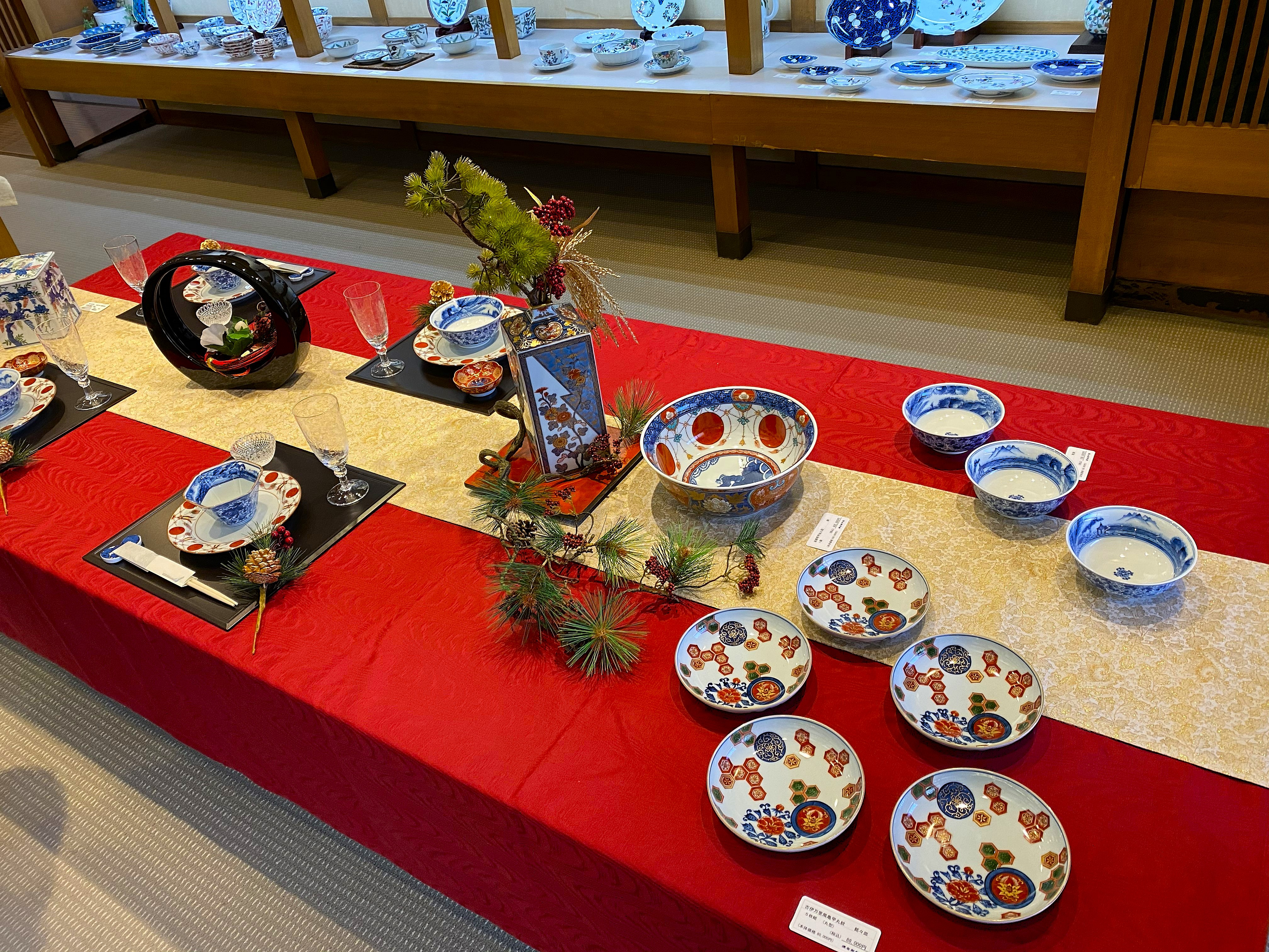 Gen-emon porcelain displayed in a formal table setting — an Imari-style table arrangement on a red cloth with pine branch decoration, blue and white landscape bowls, red and blue medallion pattern dishes, crystal glasses, and a signed Gen-emon piece as centerpiece