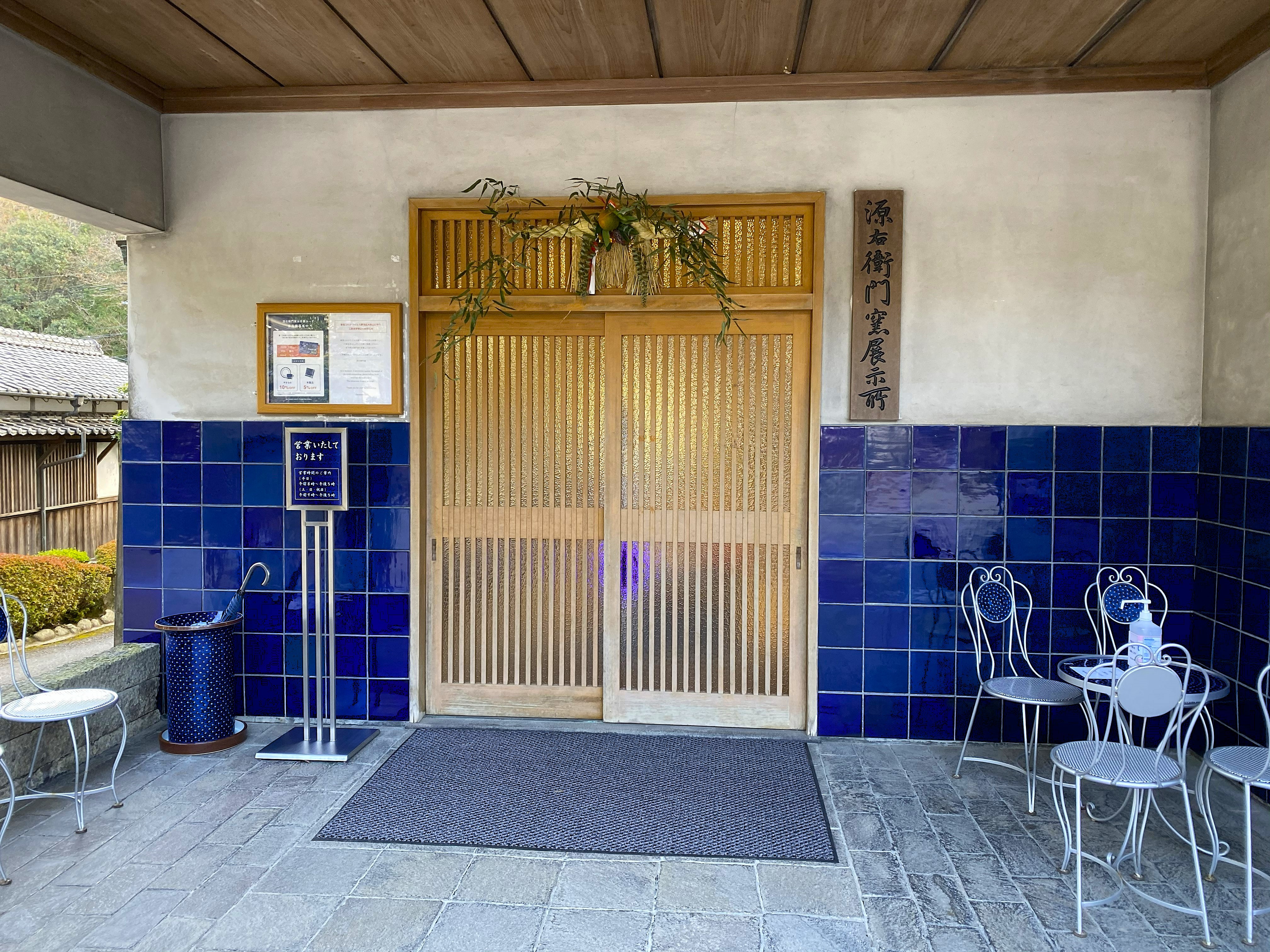 The entrance to the Gen-emon kiln showroom — wooden sliding doors with a kadomatsu pine arrangement above, a wooden sign reading 源右衛門窯展示所, cobalt blue tile walls on either side, small iron chairs and tables outside