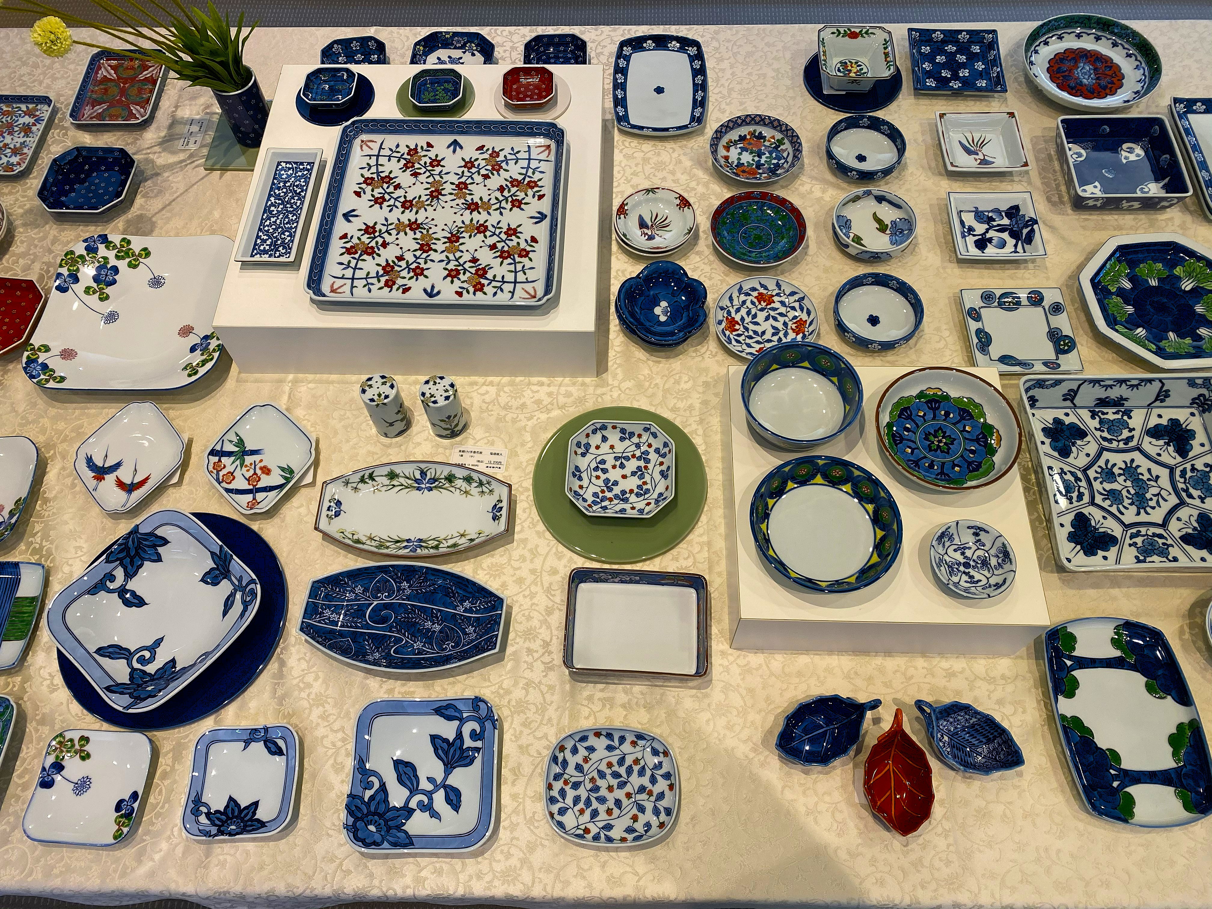 The Gen-emon showroom floor — a white tablecloth display of dozens of Gen-emon pieces: square and rectangular trays, round plates, small bowls, and individual pieces in the bold blue-white-red Gen-emon palette, some with birds and florals, others with geometric patterns