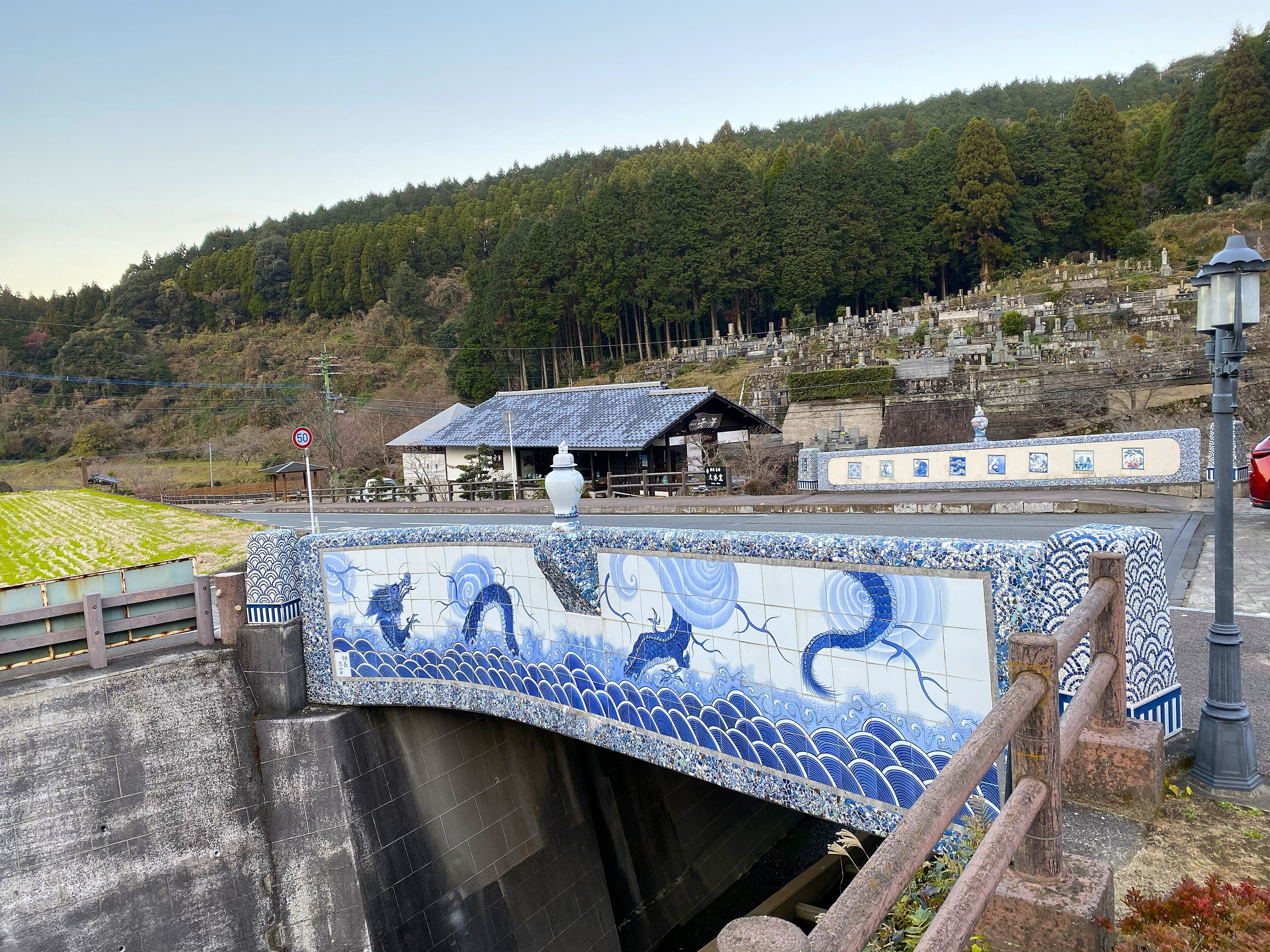 A bridge in Imari city decorated entirely with blue and white porcelain tile panels — a large dragon depicted over crashing waves on the main parapet, the mosaic tiles extending along the full length of the bridge, a porcelain vase serving as a bridge post cap, the river below and wooded hillside behind