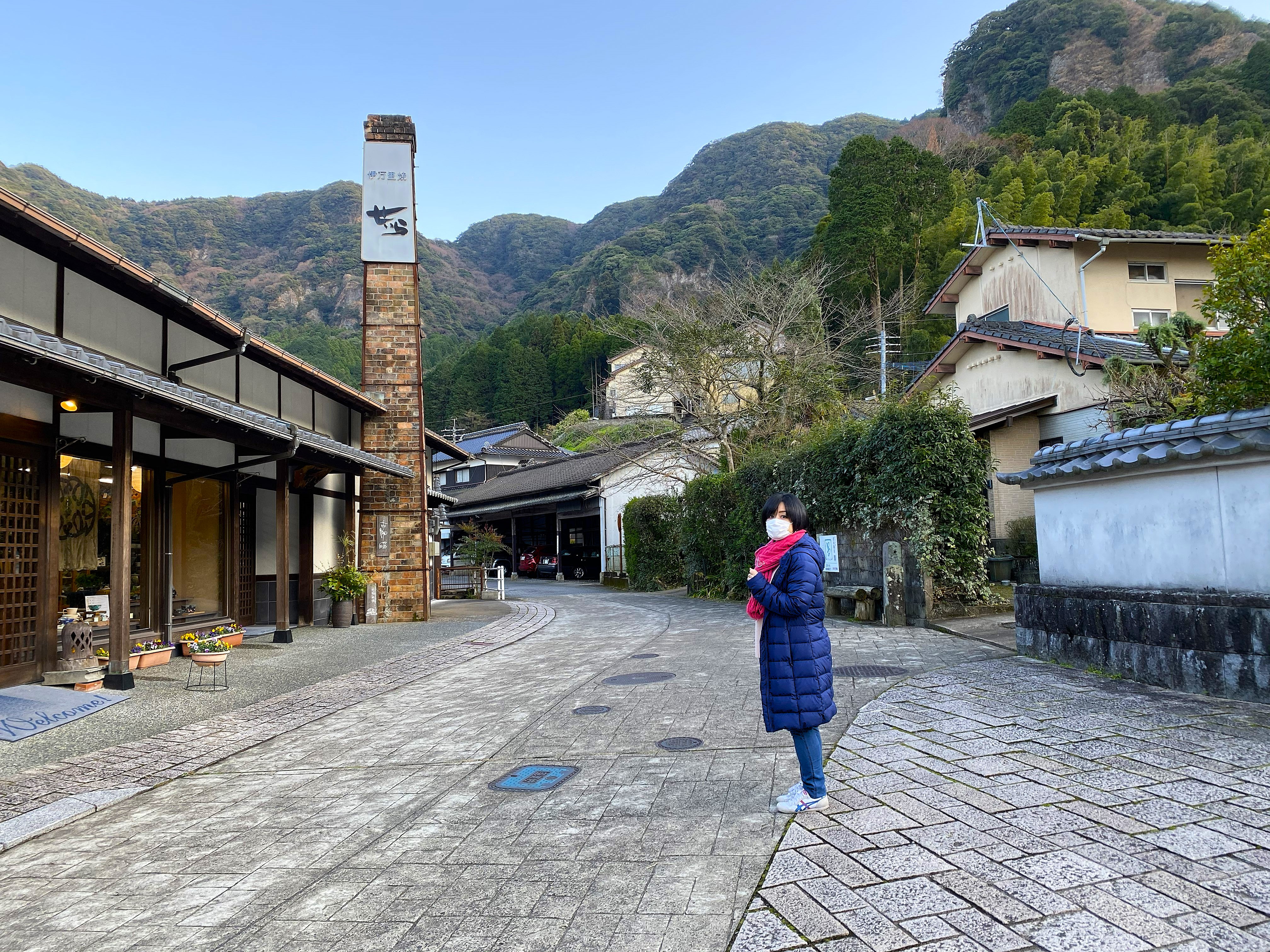 Ayumi standing on a quiet stone-paved street in Imari's pottery district — a tall brick kiln chimney rises behind her on the left with a sign reading 御写意城, traditional townhouse buildings line the street, mountains in the background under a clear blue sky