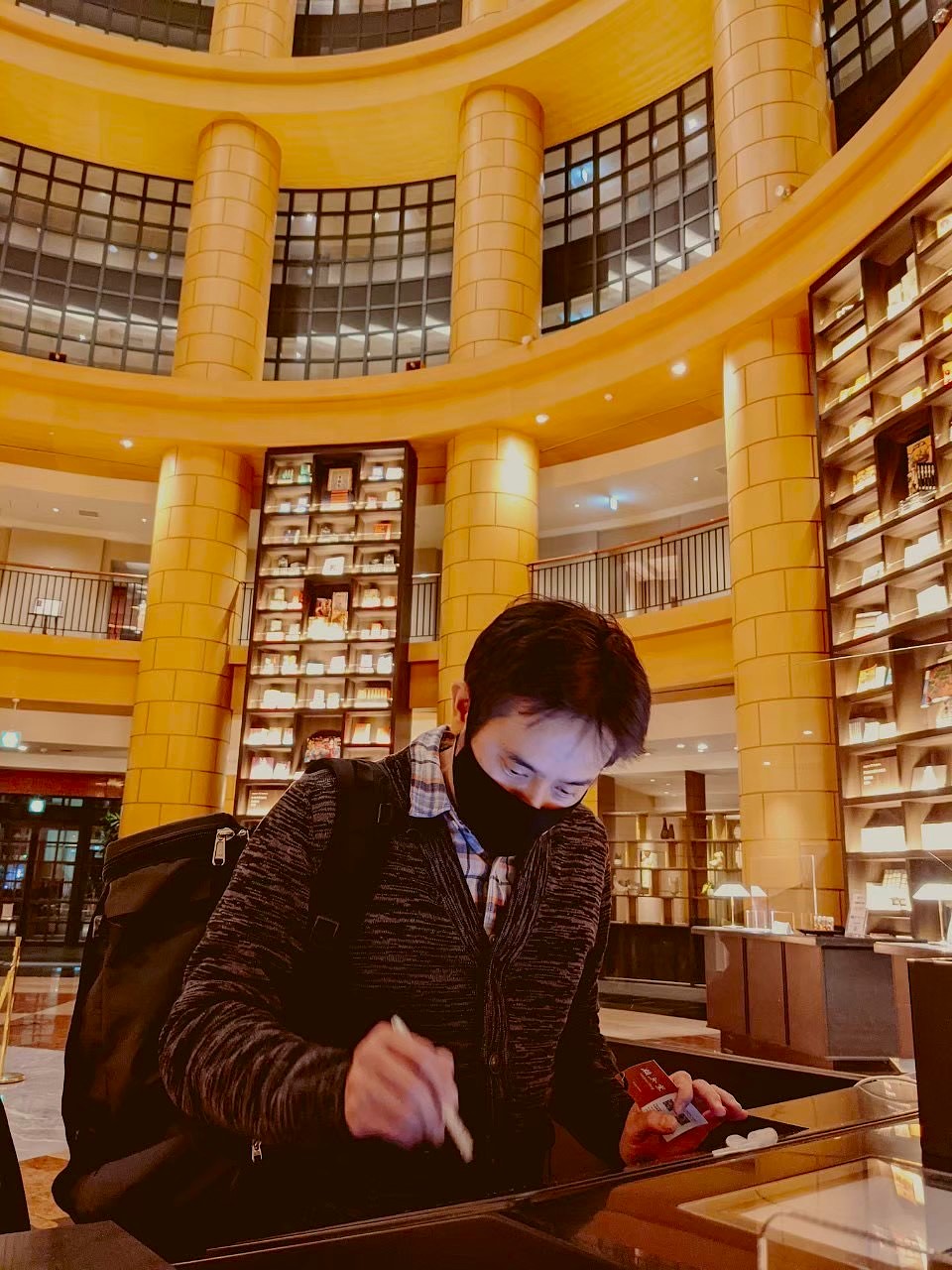 John checking in at The Basics Fukuoka hotel — the dramatic rotunda lobby rises behind him with warm amber columns and floor-to-ceiling bookshelves on multiple levels, the hotel's signature library-themed design immediately visible