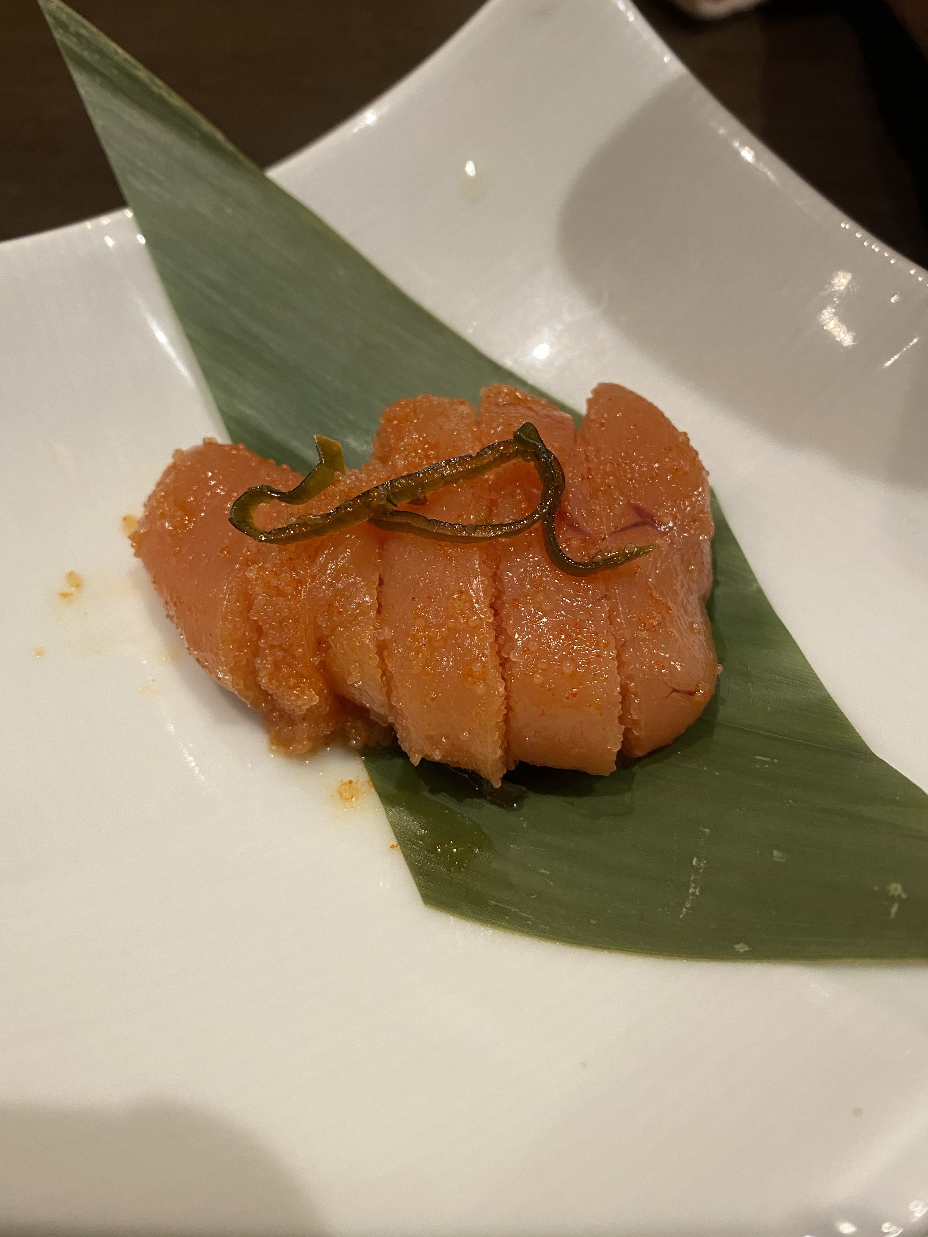 A piece of Fukuoka mentaiko — karashi mentaiko, spicy marinated pollock roe — sliced and presented on a bamboo leaf with a thin kelp garnish, on a white plate